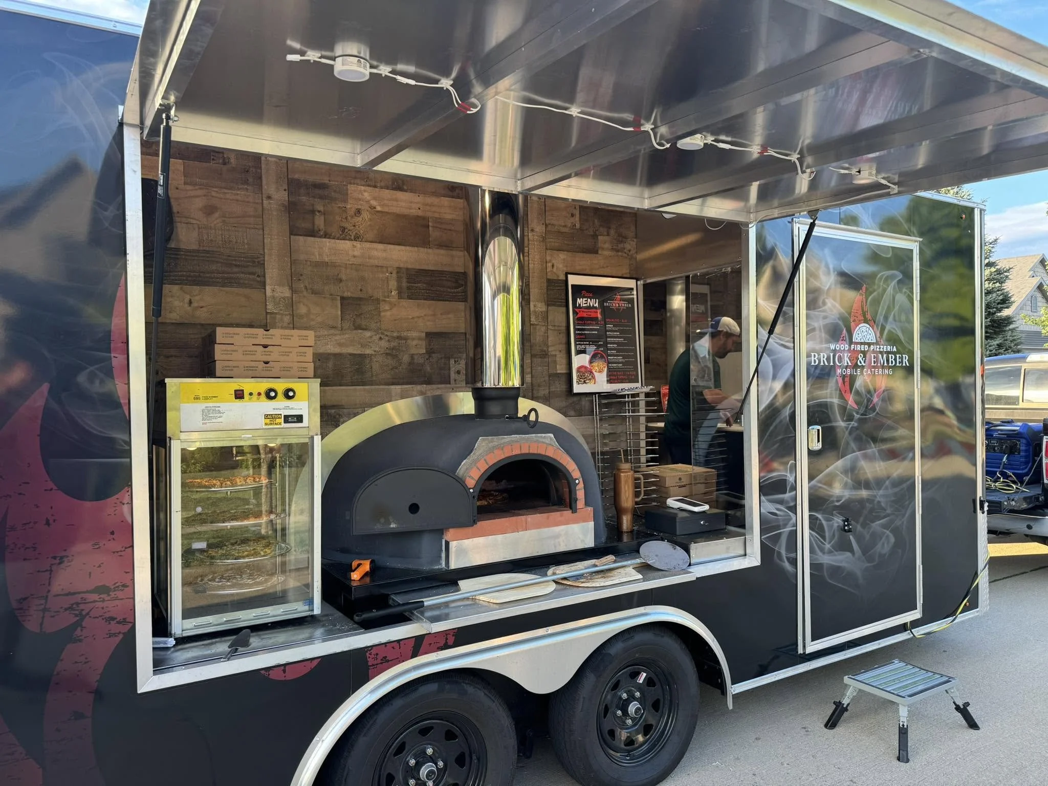 A food truck named Brick & Ember Wood Fired Pizzeria with a pizza oven and menu displayed inside, set up outdoors during daytime.