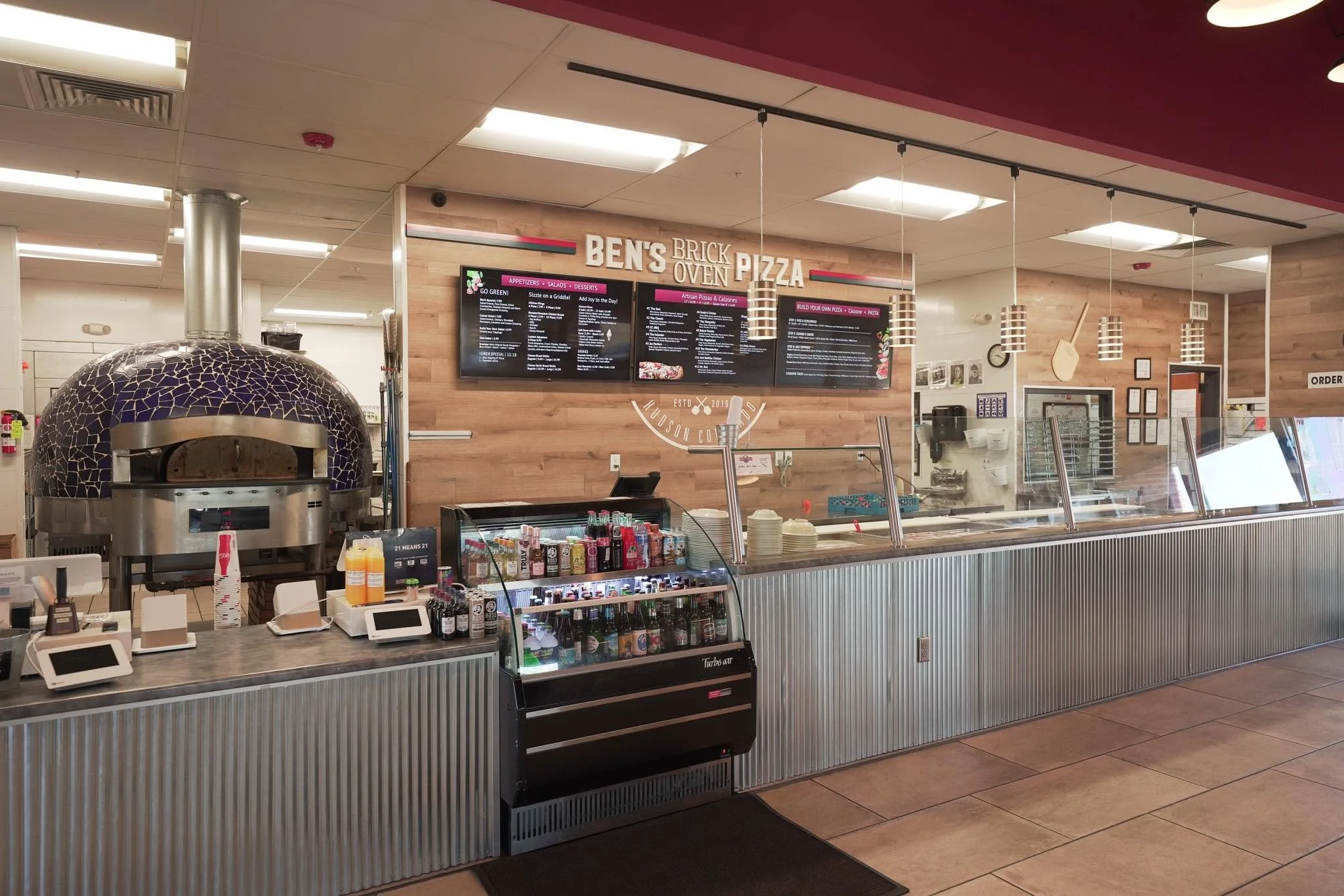 A pizza counter at Ben's Brick Oven Pizza with a pizza oven, menu screens, drink cooler, and order area.