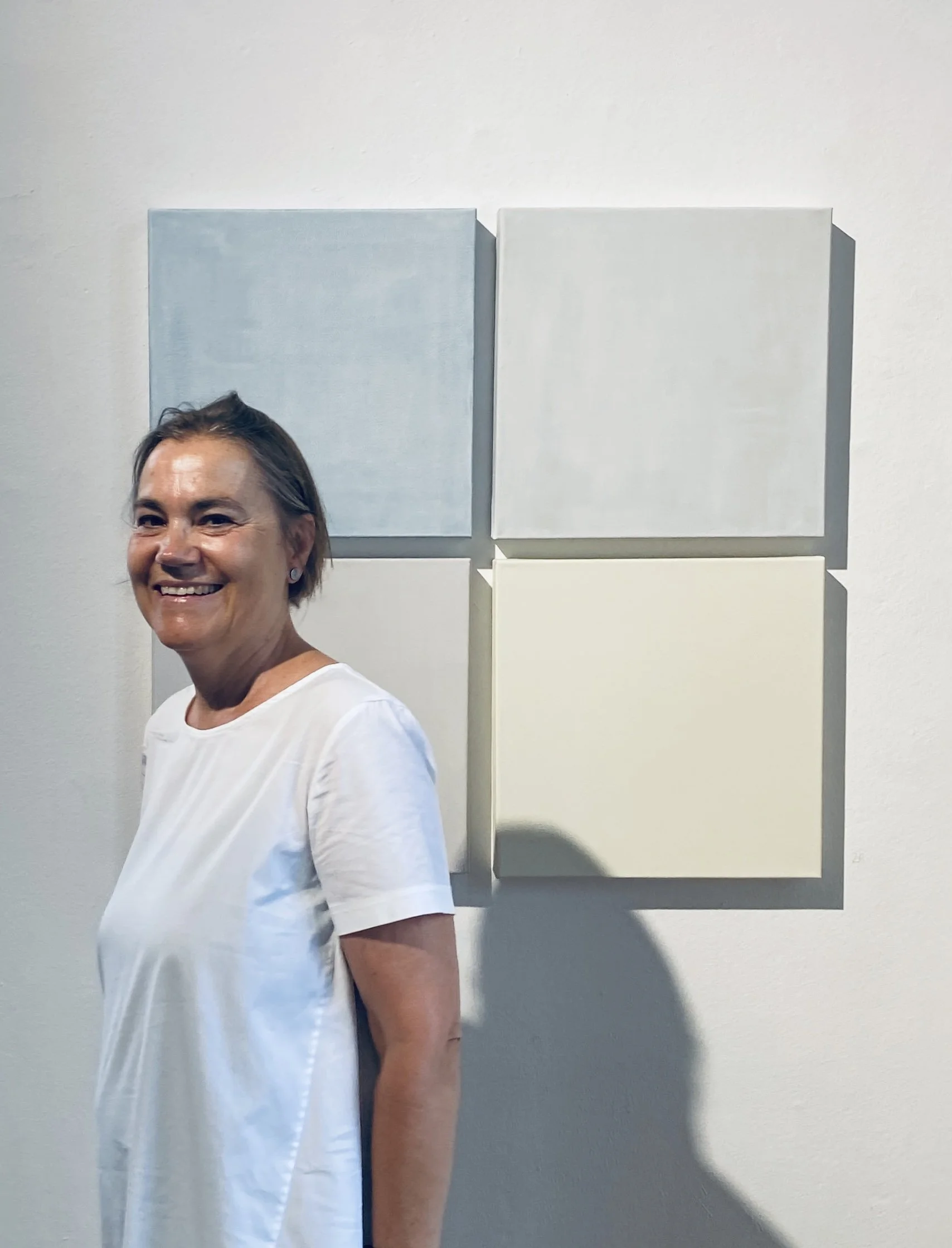 A woman smiling in front of four blank square canvases mounted on a wall, with three on the top row and one on the bottom row.