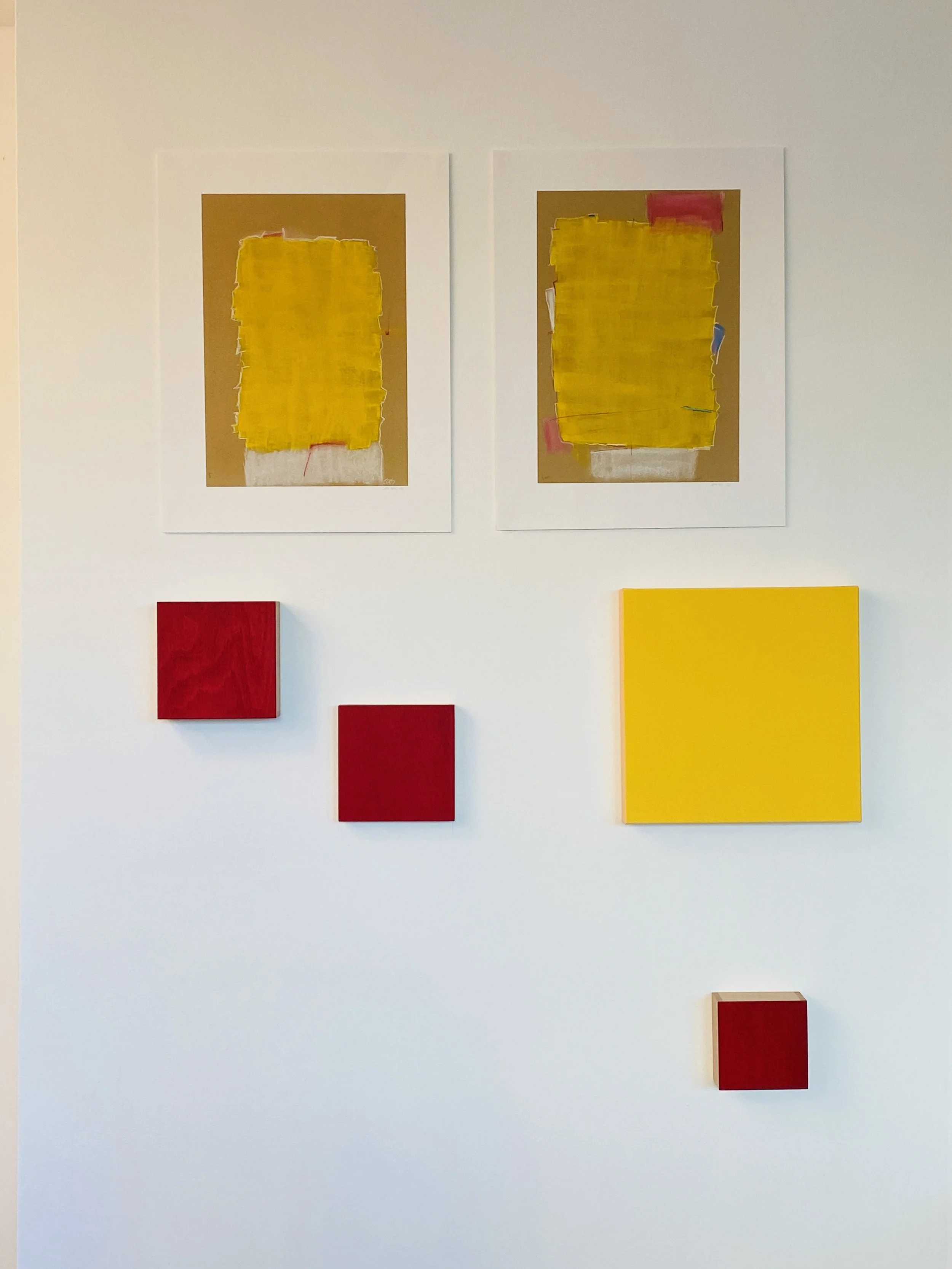 Two abstract paintings featuring large yellow rectangular shapes on brown backgrounds, along with smaller red and yellow canvases on a white wall.