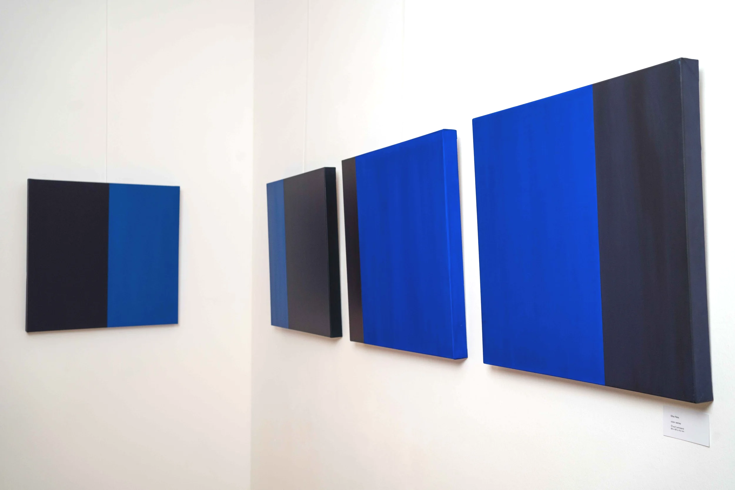Vertical abstract paintings in shades of blue and black, hanging on a white gallery wall.