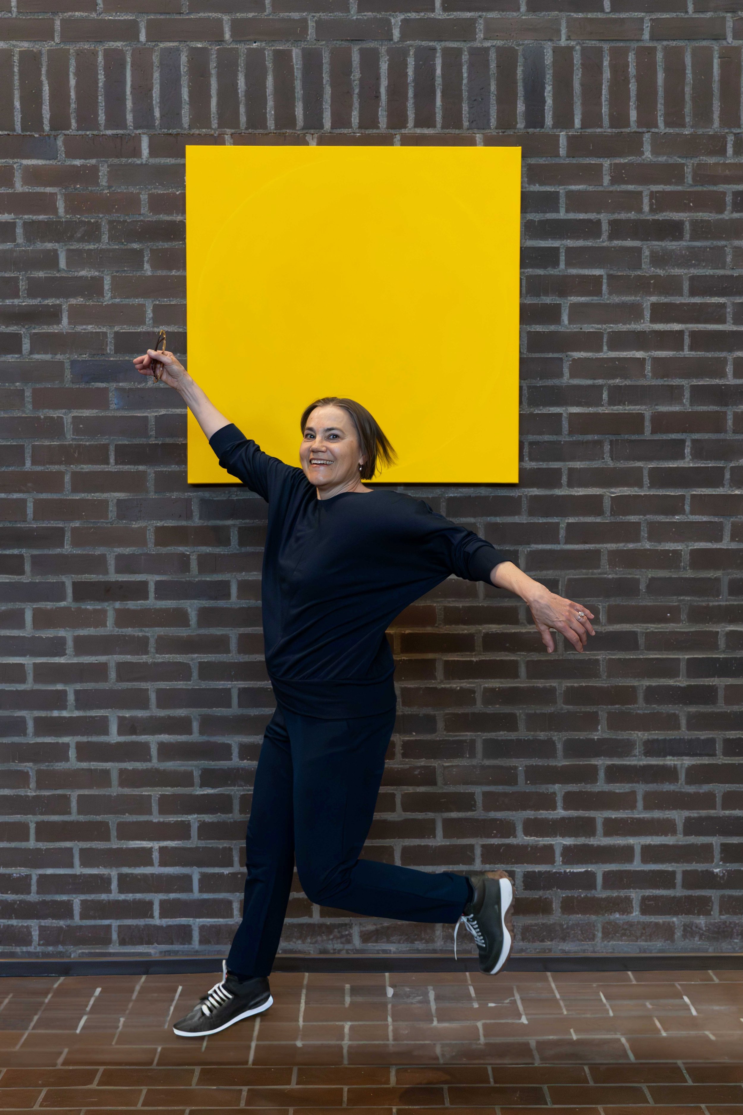 A woman in black athletic clothing and sneakers strikes a dance pose against a brick wall with a large yellow square art piece. She has short hair and smiles while extending her right arm up and her left arm to the side, with her left leg bent and raised.