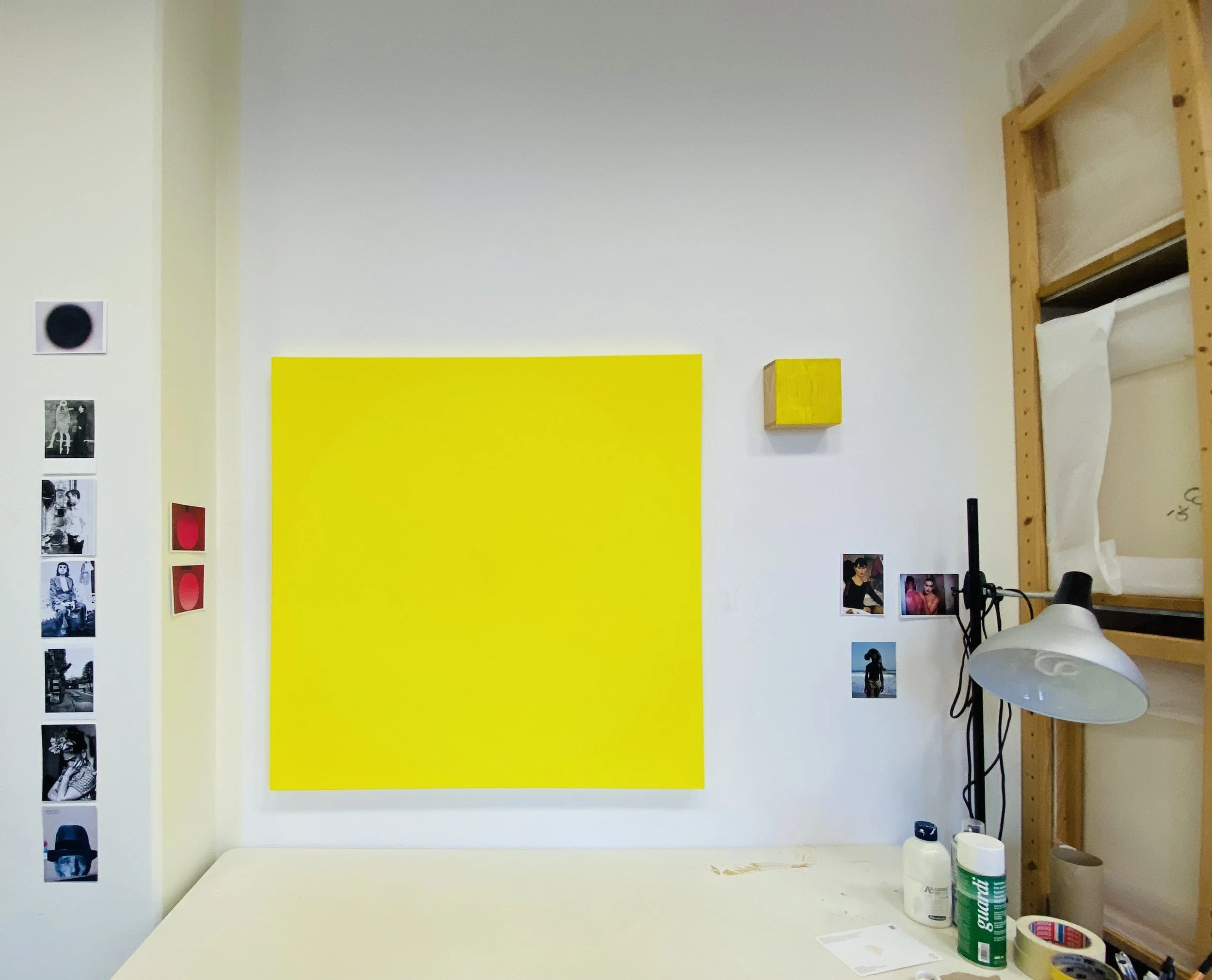 Interior of an art gallery or studio with a white wall displaying a large yellow painting or poster. To the right, a wooden shelving unit holds paper or canvas and other supplies. On the left wall, black and white photographs are displayed vertically. The bottom of the image shows a white table with art supplies, including a green spray can, a white container, and other small items.