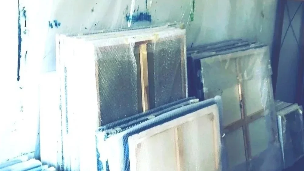 Several large window screens stacked on a wall, with some covered in plastic or foam, in a workshop or construction site.