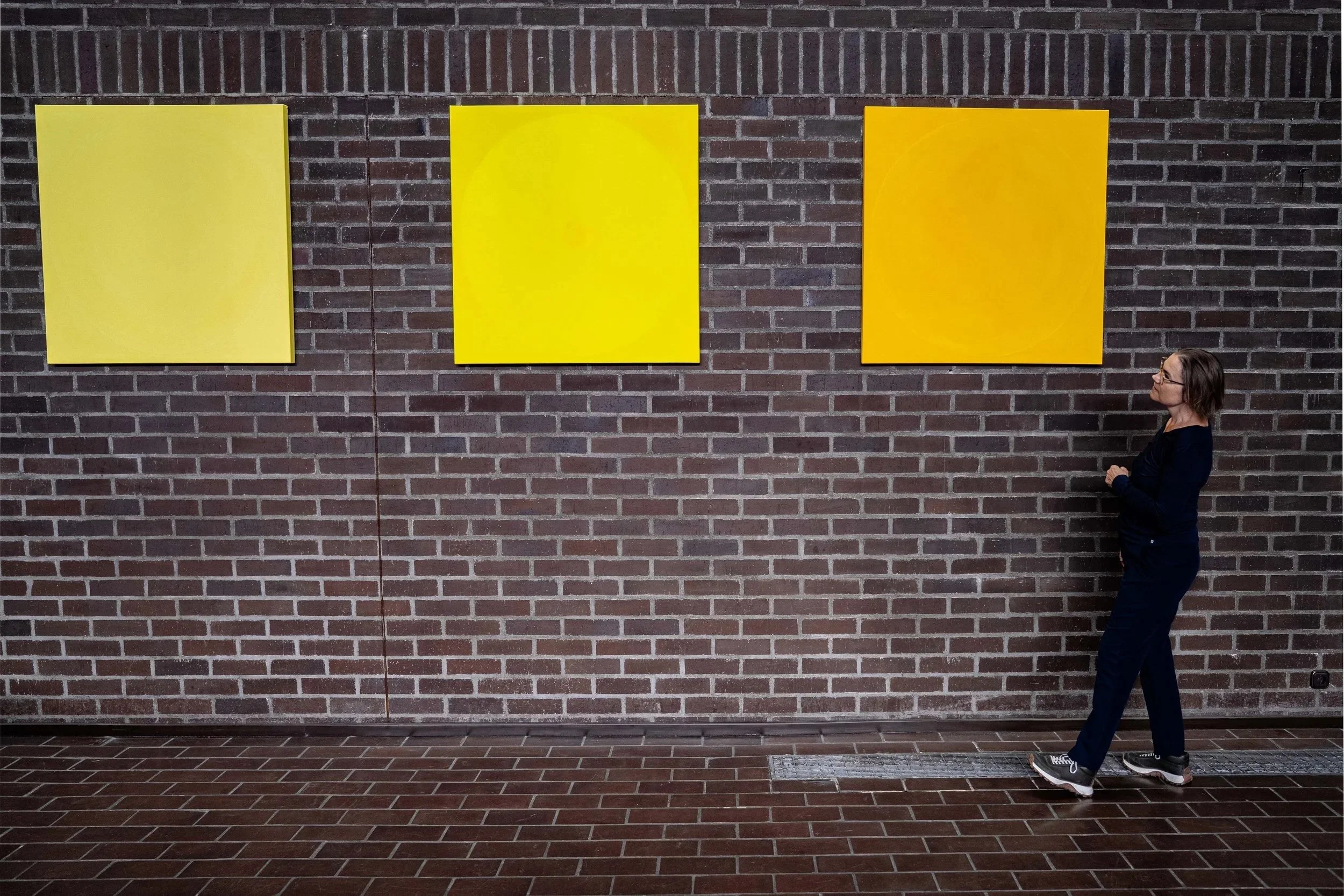 A woman with dark hair and glasses stands in front of a brick wall with three large yellow abstract paintings. She is wearing a dark top, dark pants, and sneakers, and is looking to her right.