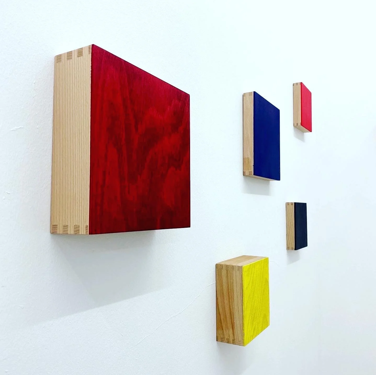 Wall-mounted art pieces in various colors, including red, blue, black, yellow, and natural wood, arranged in an alternating pattern on a white wall.