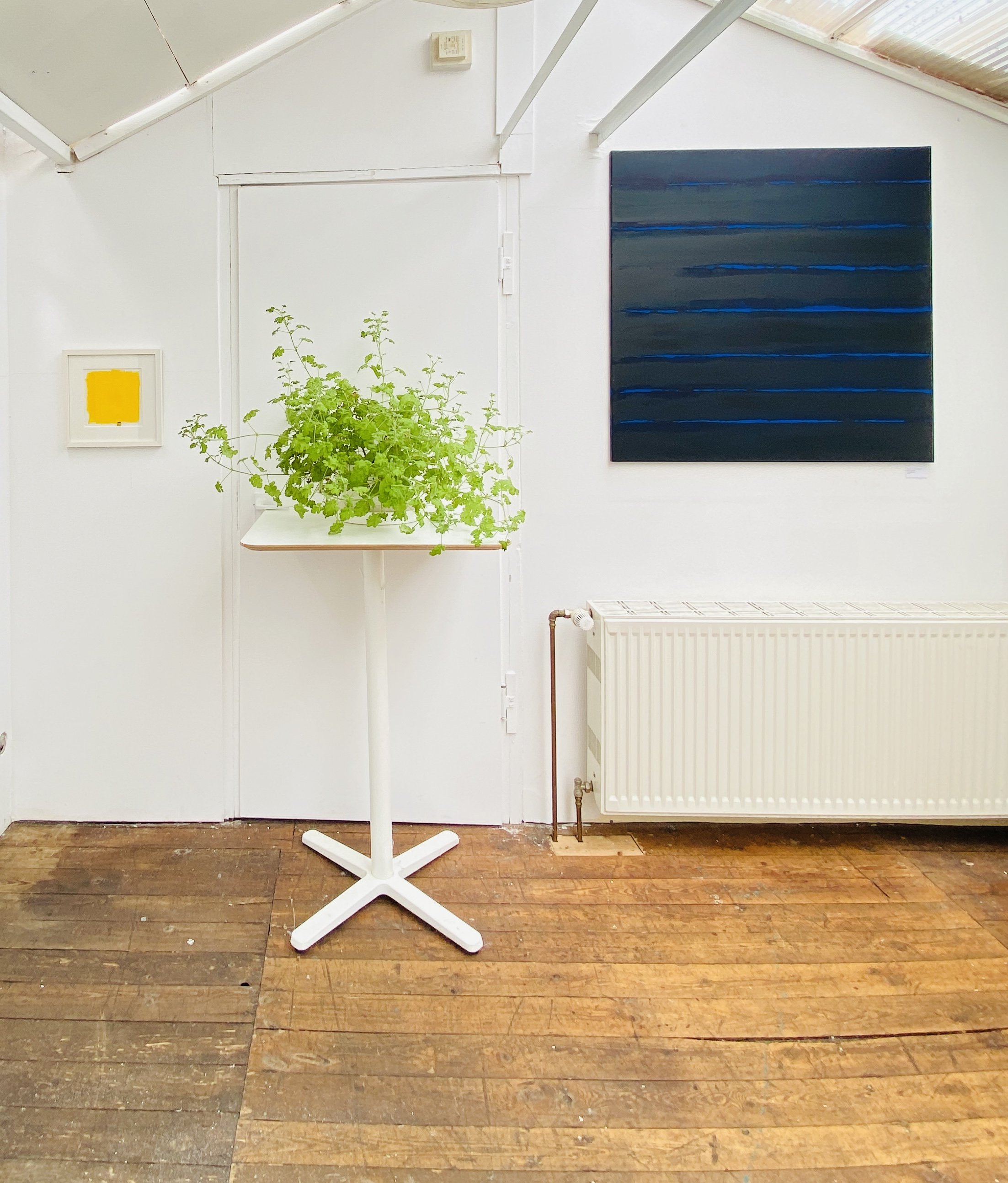 A white stand with a plant on it, positioned in front of a white wall with two framed pieces of art: a small square yellow piece and a large rectangular dark abstract piece. The floor is wooden with visible wear and tear.