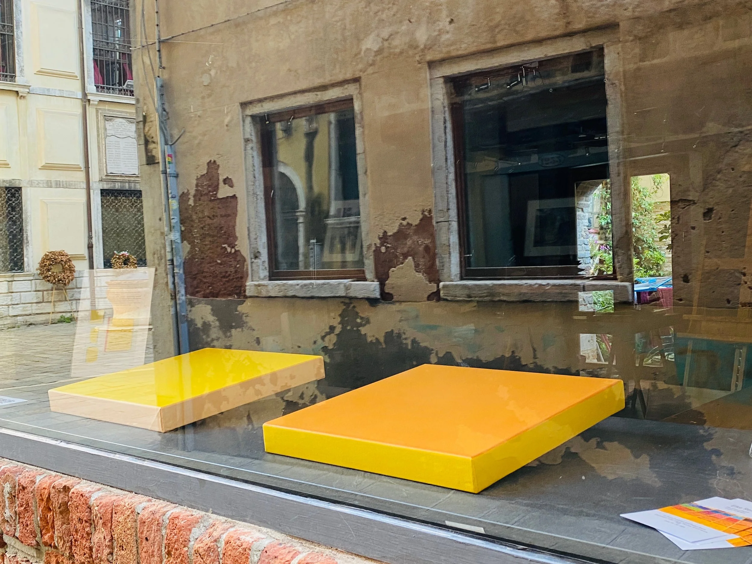 View of a building's exterior with worn yellow facade and a window, seen through a shop window with yellow tables inside.