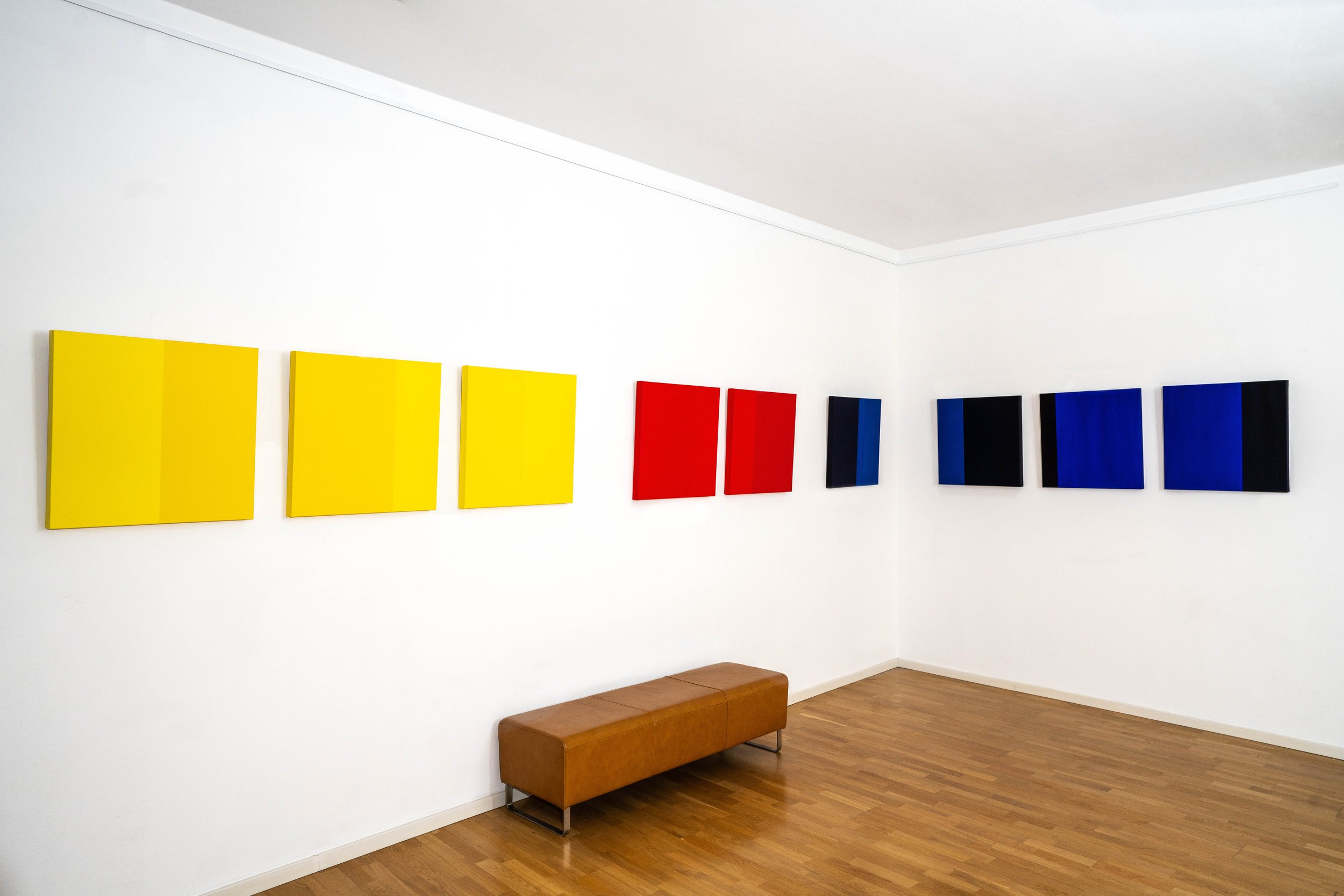 Gallery wall with abstract paintings in yellow, red, blue, and black on a white wall, with a wooden bench below.