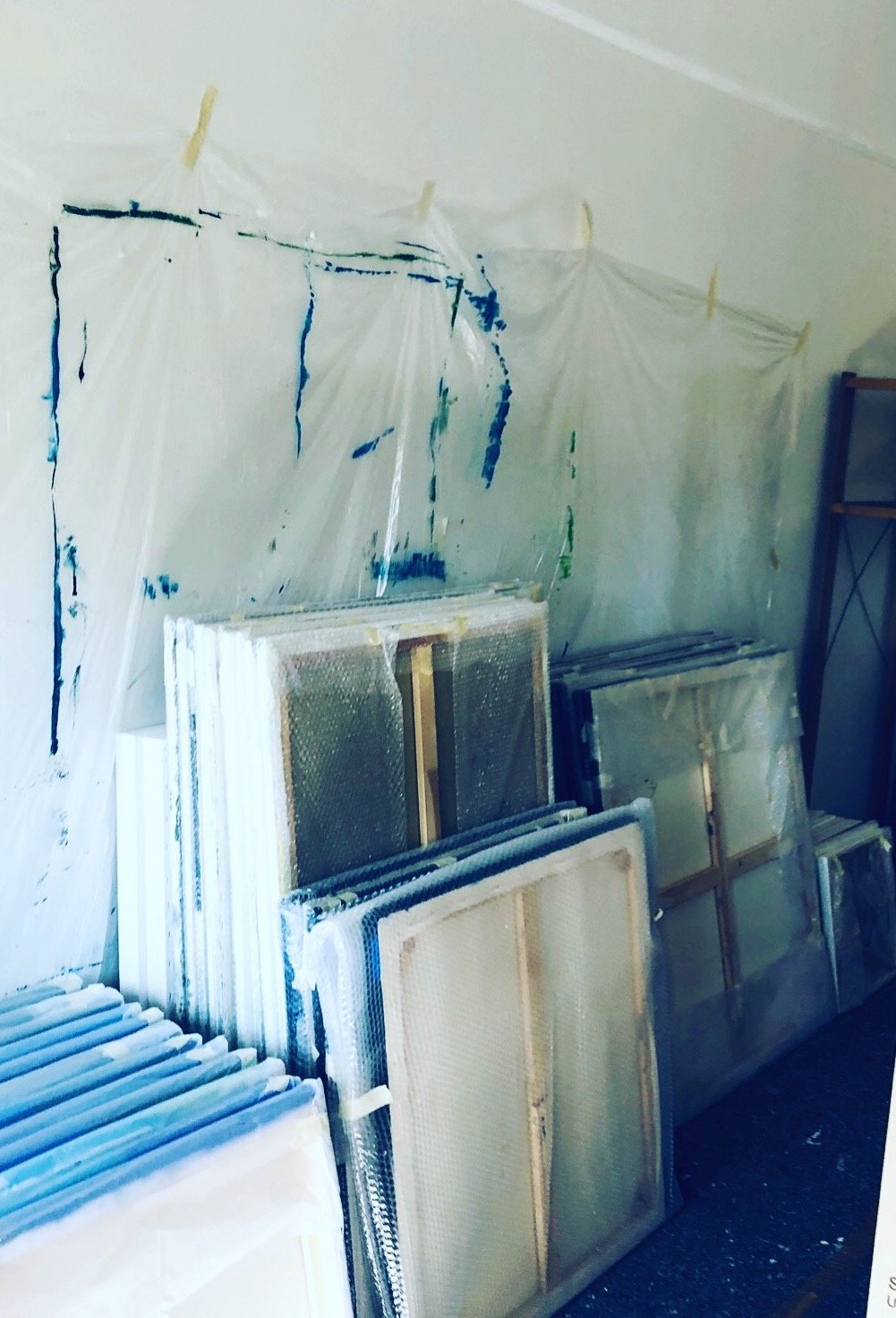A wall with multiple window screens leaning against it, some wrapped in plastic, and a plastic sheet with clothespins hanging above, secured to a wire.