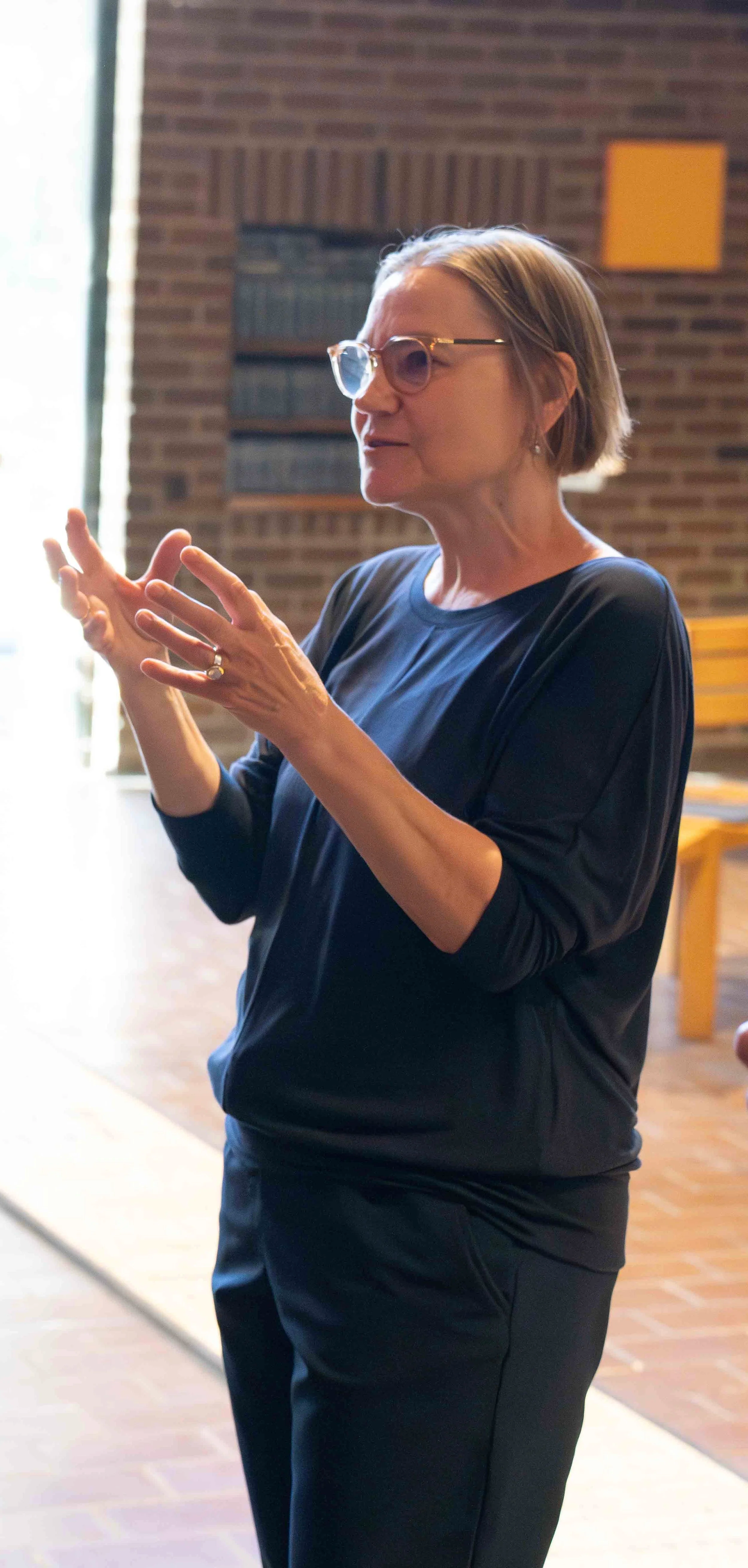 A woman with short hair, glasses, and earrings is speaking or explaining something inside a room with brick walls. She is wearing a black top and black pants.