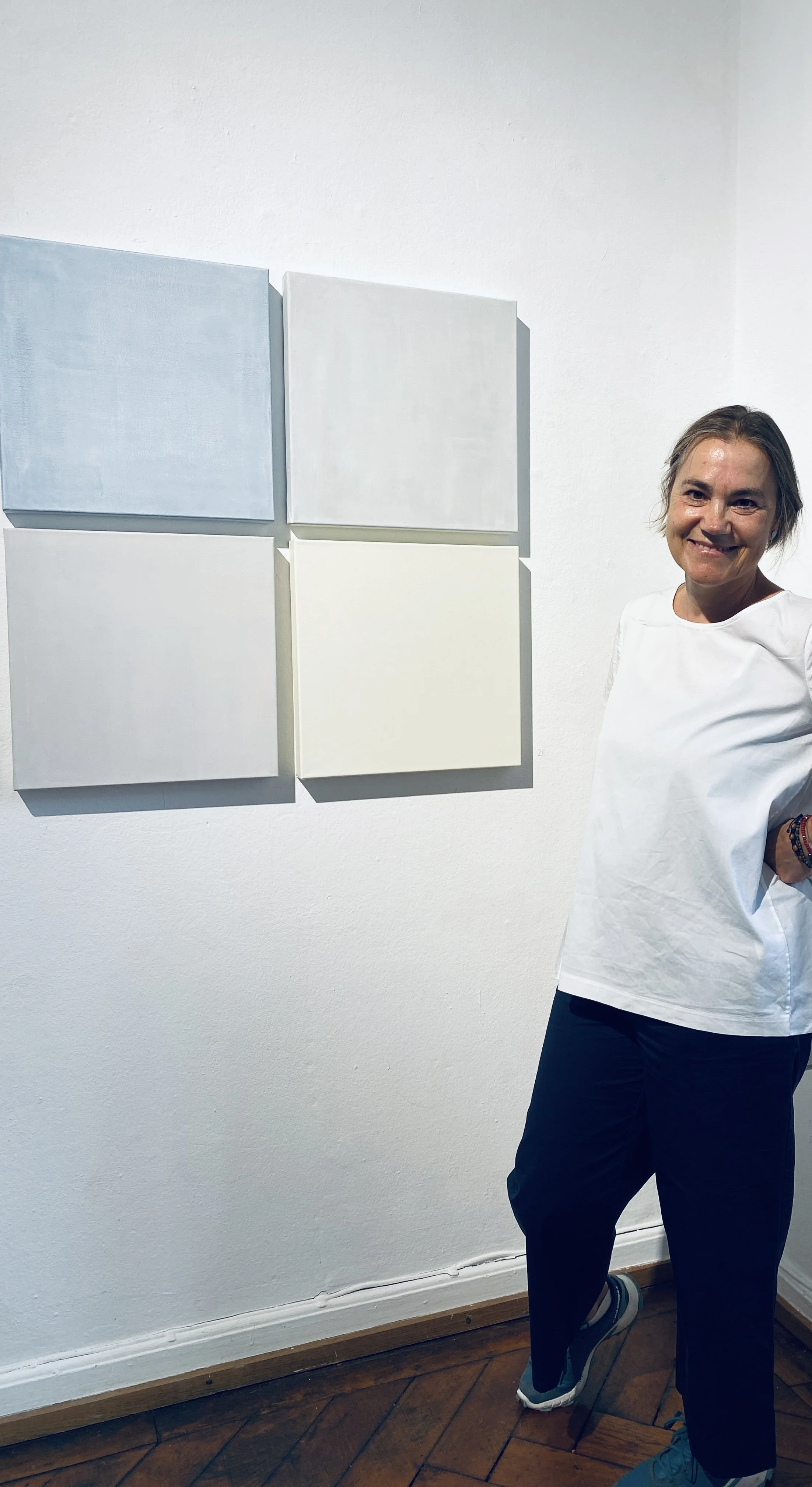 A woman standing in front of four square abstract paintings on white wall, wearing a white top and black pants, in an art gallery.
