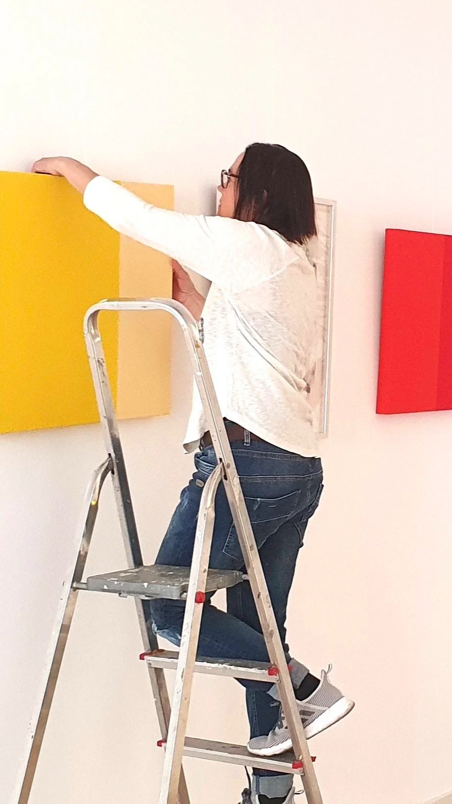 A woman on a step ladder arranging or hanging colorful squares on a white gallery wall.