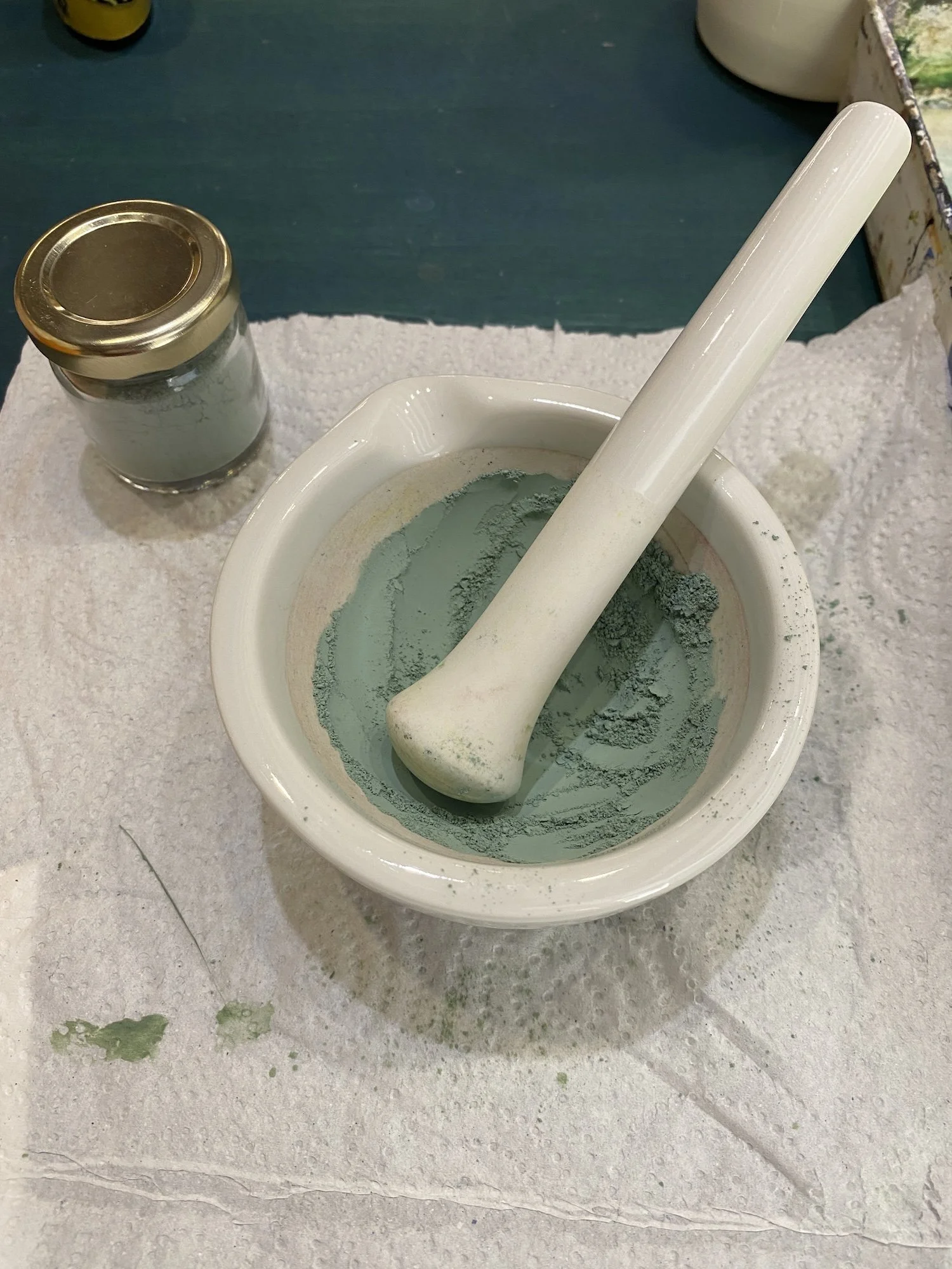 A mortar and pestle with green powder, a small jar with a white substance, and a paper towel on a green work surface.