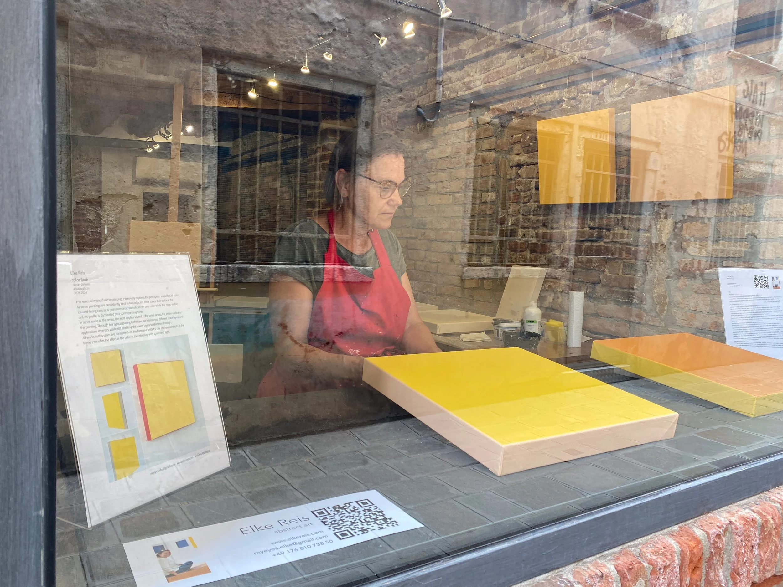 View of a person with glasses and a red apron working in an art studio with exposed brick walls, seen through a window with informational posters and QR code in the foreground.