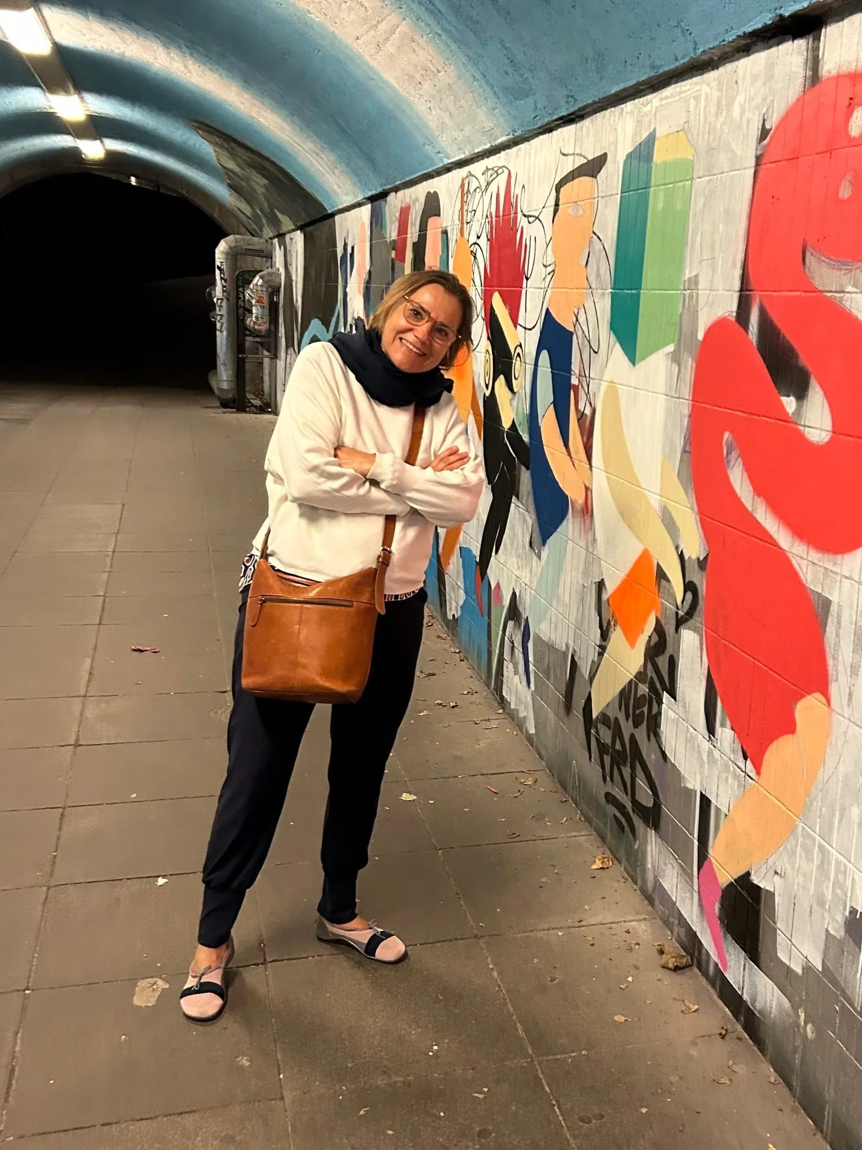 A woman with glasses, a beige jacket, black pants, and slippers, standing in front of colorful graffiti art on a wall in an underground tunnel.