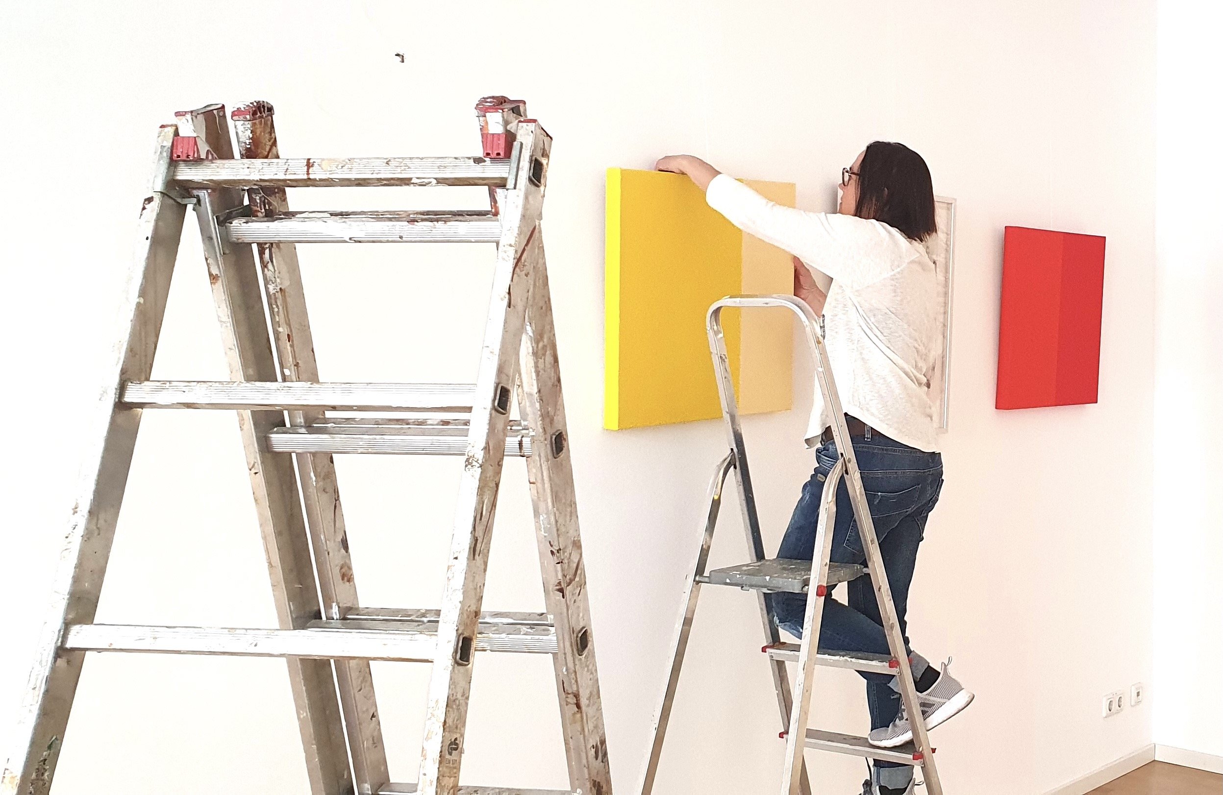 A woman with dark hair and tie-dye jeans is standing on a step ladder, reaching up to a yellow canvas on a white gallery wall. There is a red canvas hanging on the wall behind her, and a wooden stepladder is in the foreground.