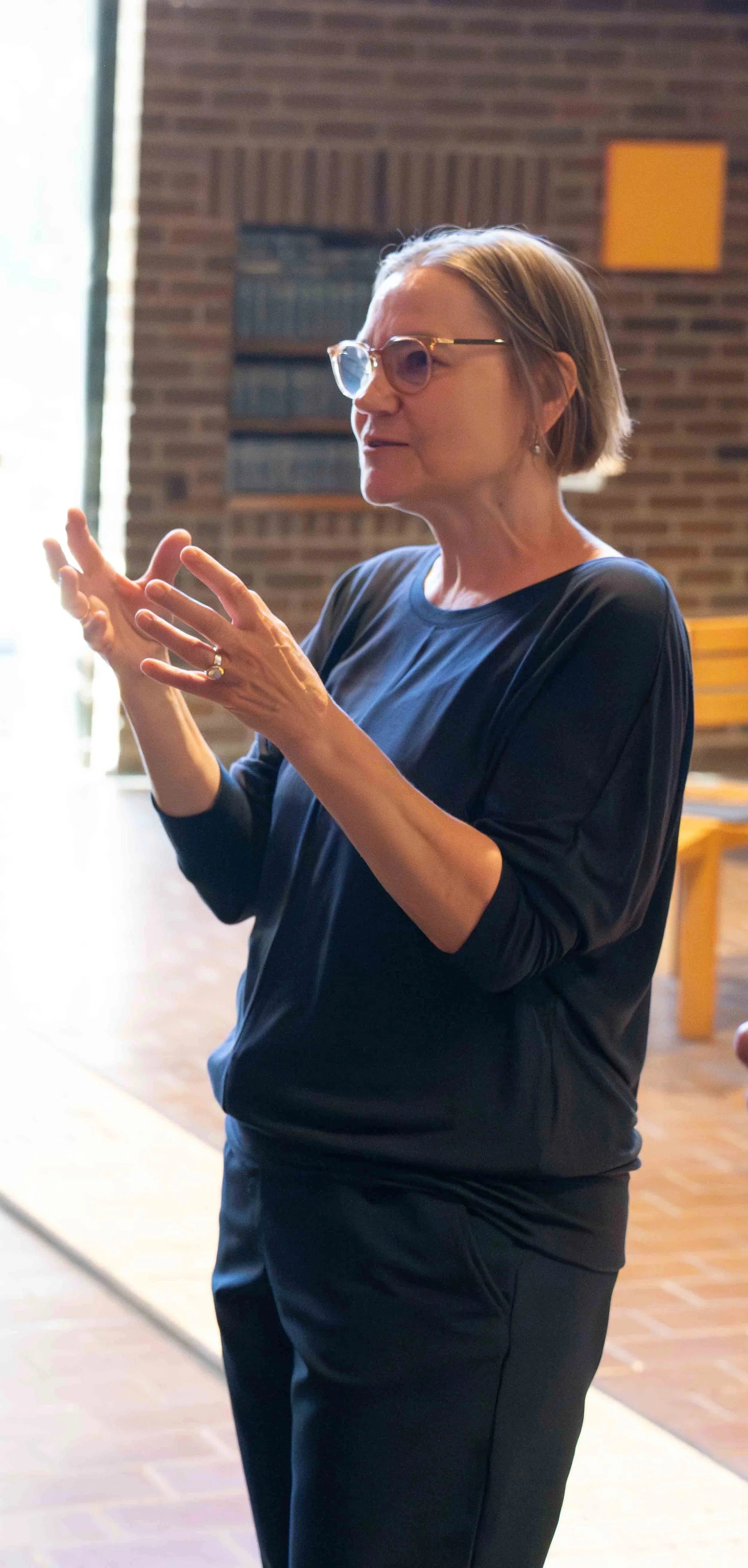 A woman with short hair, sunglasses, and earrings is speaking or explaining with her hands in a room with brick walls and wooden furniture.