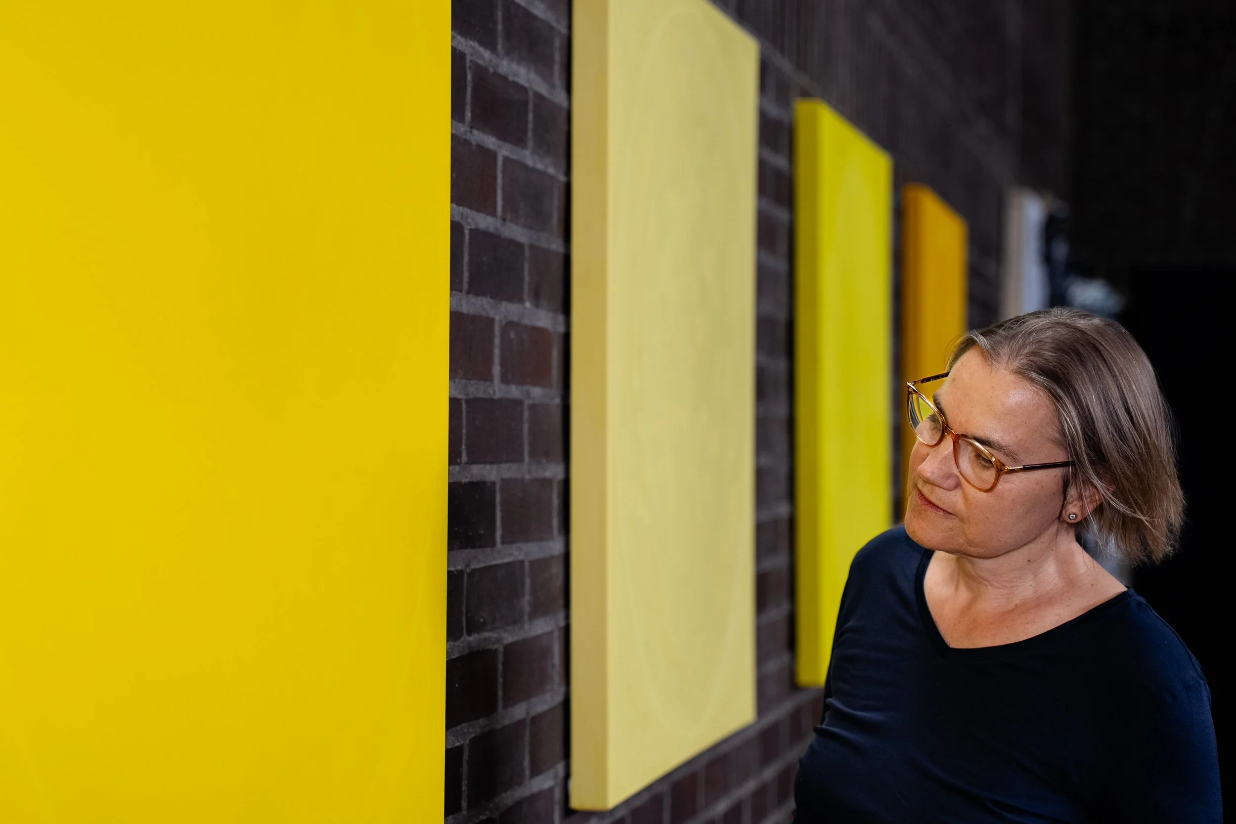 A woman with glasses and short hair looking at yellow rectangular panels mounted on a brick wall.