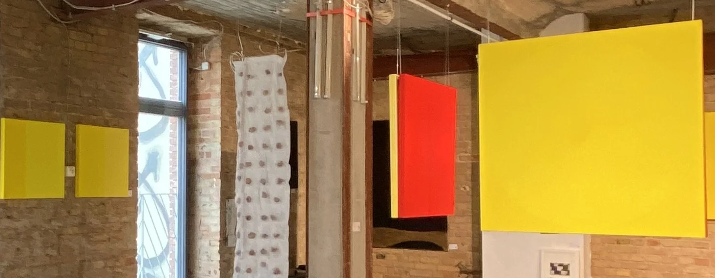 Interior of a room with exposed brick walls, colorful hanging panels in red and yellow, a white fabric with a polka dot pattern hanging near a window, and modern lighting fixtures.
