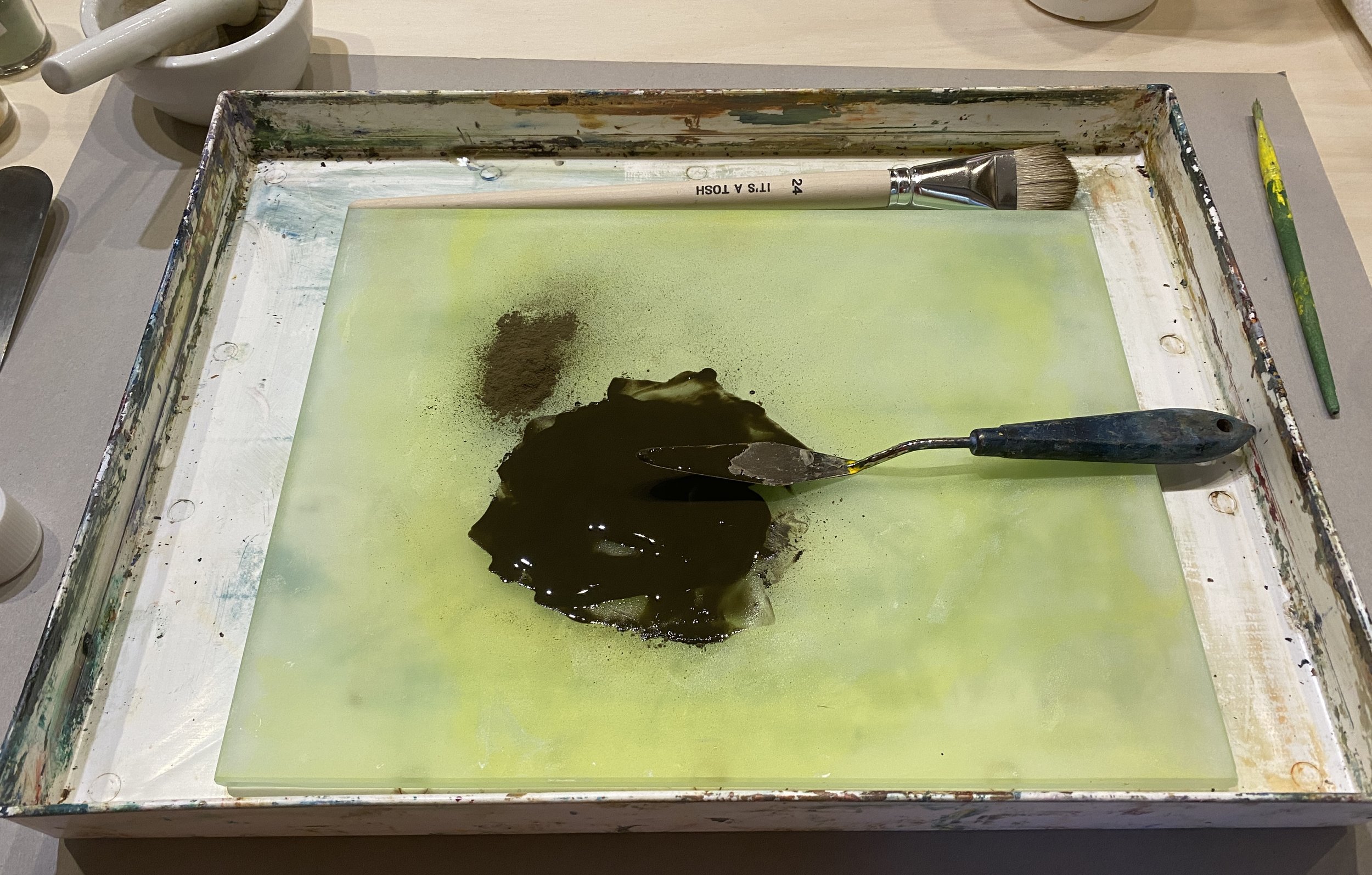 An art workspace with a canvas, dark paint, and a paintbrush resting on the canvas. The workspace is surrounded by a paint-stained tray and various art tools.