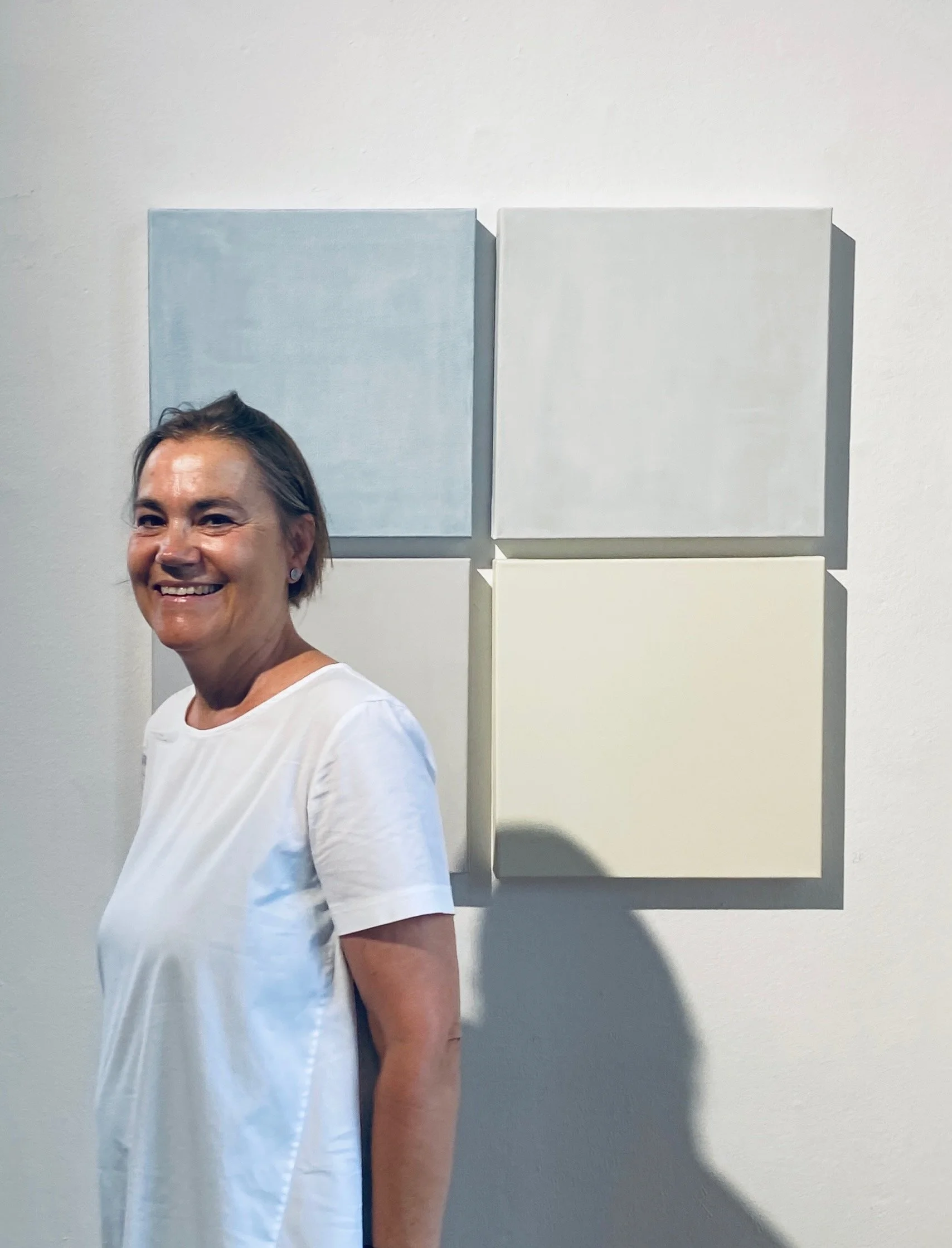 A smiling woman in a white shirt, with light brown hair, standing in front of a white wall with four abstract square paintings, including two large grey squares and two smaller cream and beige squares.