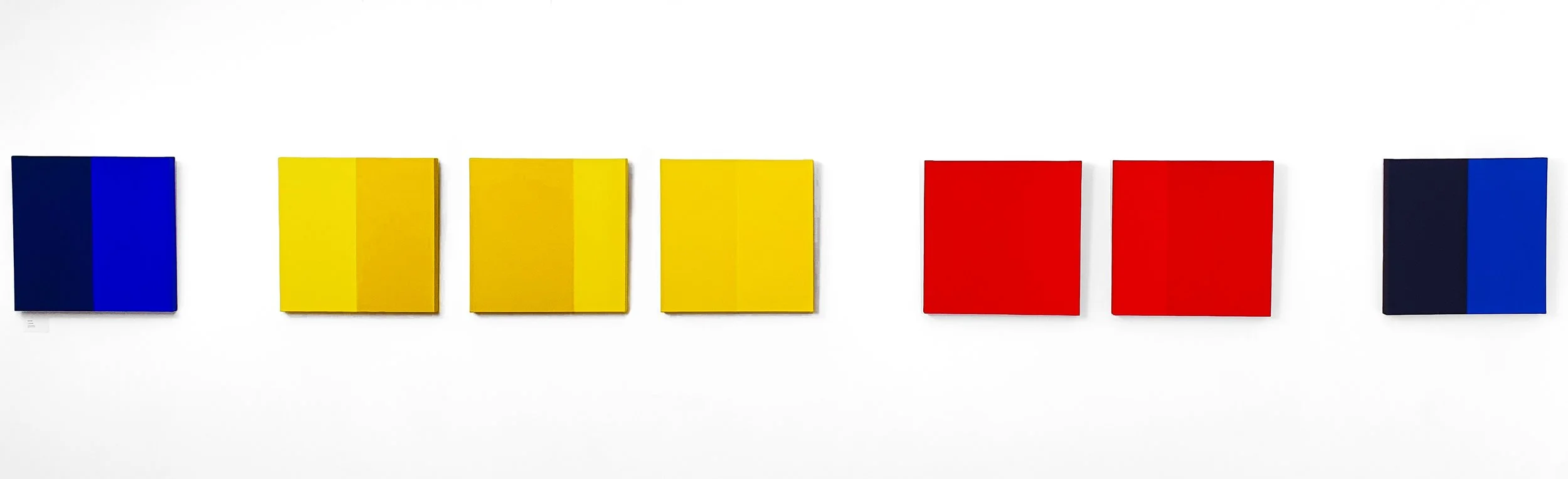 A series of seven square abstract paintings arranged in a row on a white gallery wall. The paintings feature vertical color blocks in shades of blue, yellow, and red.