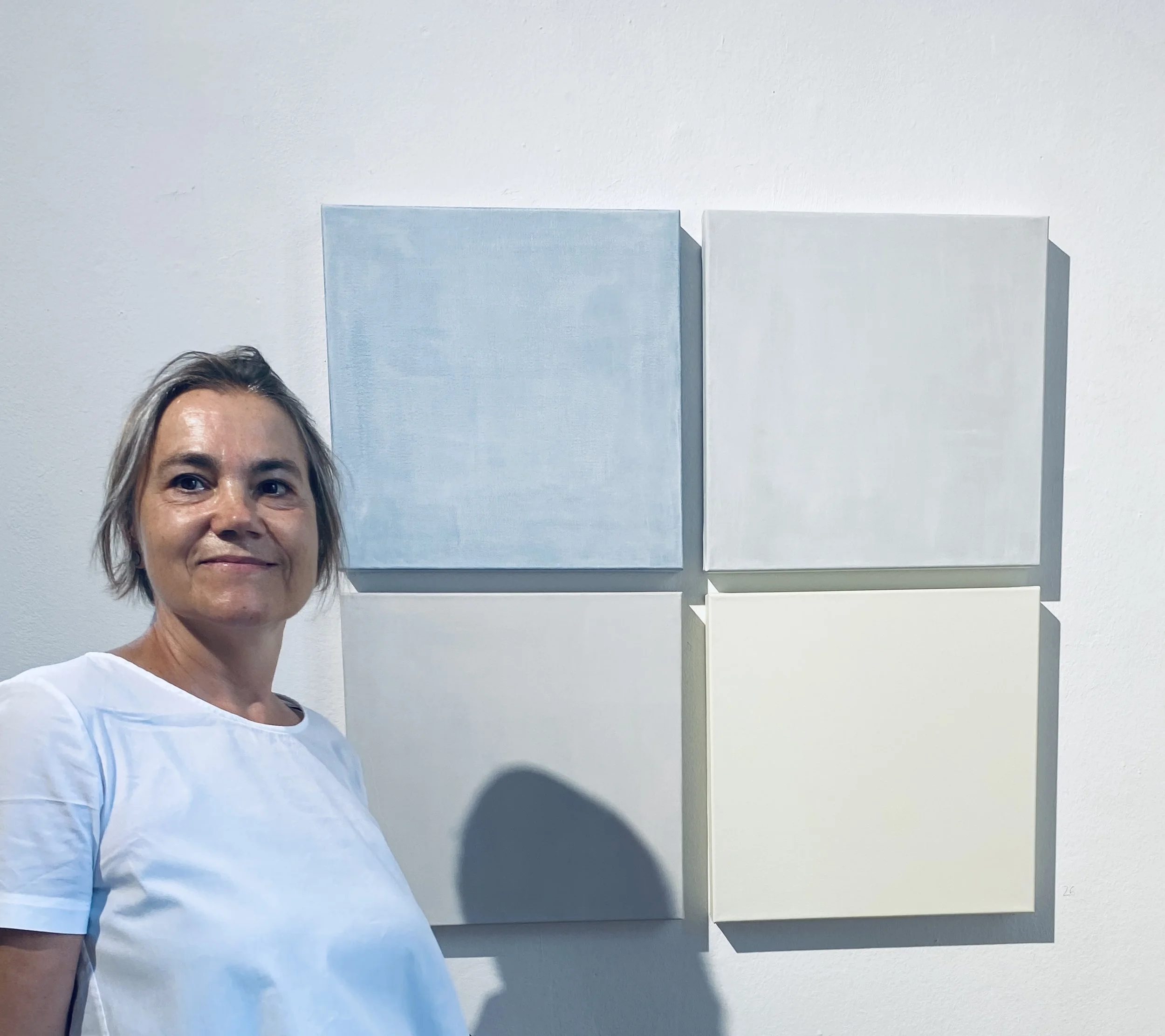 A woman with short, light-colored hair smiling at the camera, wearing a white shirt, standing in front of a white wall adorned with several abstract paintings with soft pastel colors.
