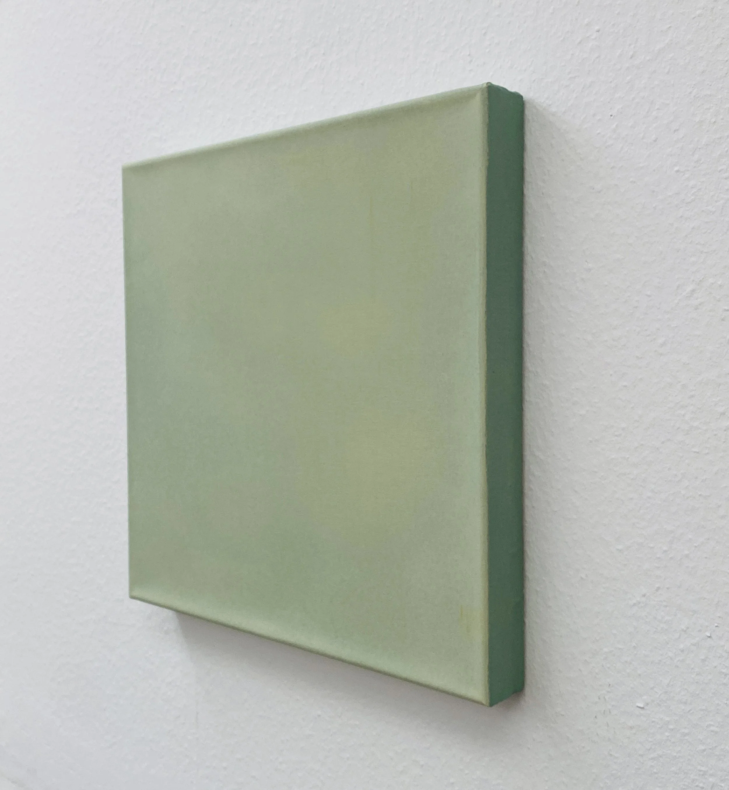 Close-up of a rectangular canvas with a light green front and darker green edges, mounted on a white textured wall.