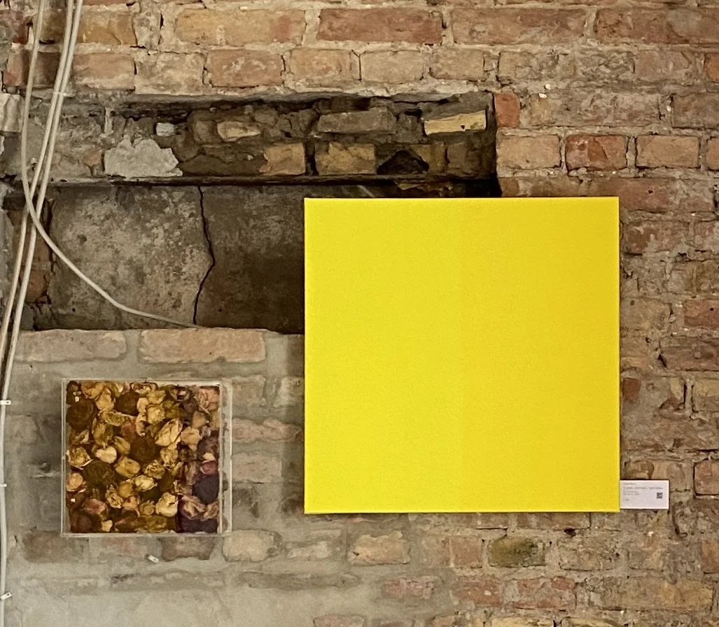 An art installation with a large, solid yellow square canvas and a smaller collage of dried flowers, all mounted on an exposed brick wall.
