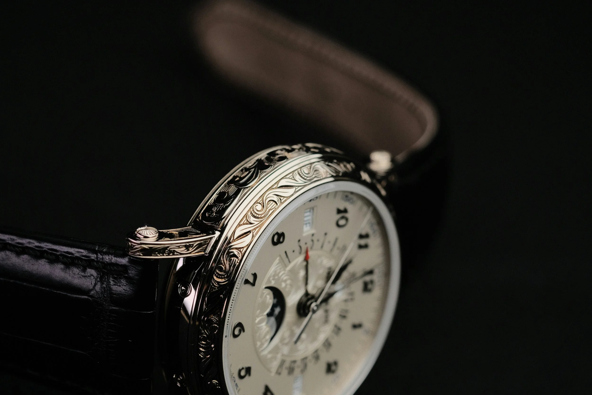 Nahaufnahme einer eleganten Armbanduhr mit silbernem Zifferblatt, schwarzen Ziffern und einem schwarzen Lederarmband, vor dunklem Hintergrund.