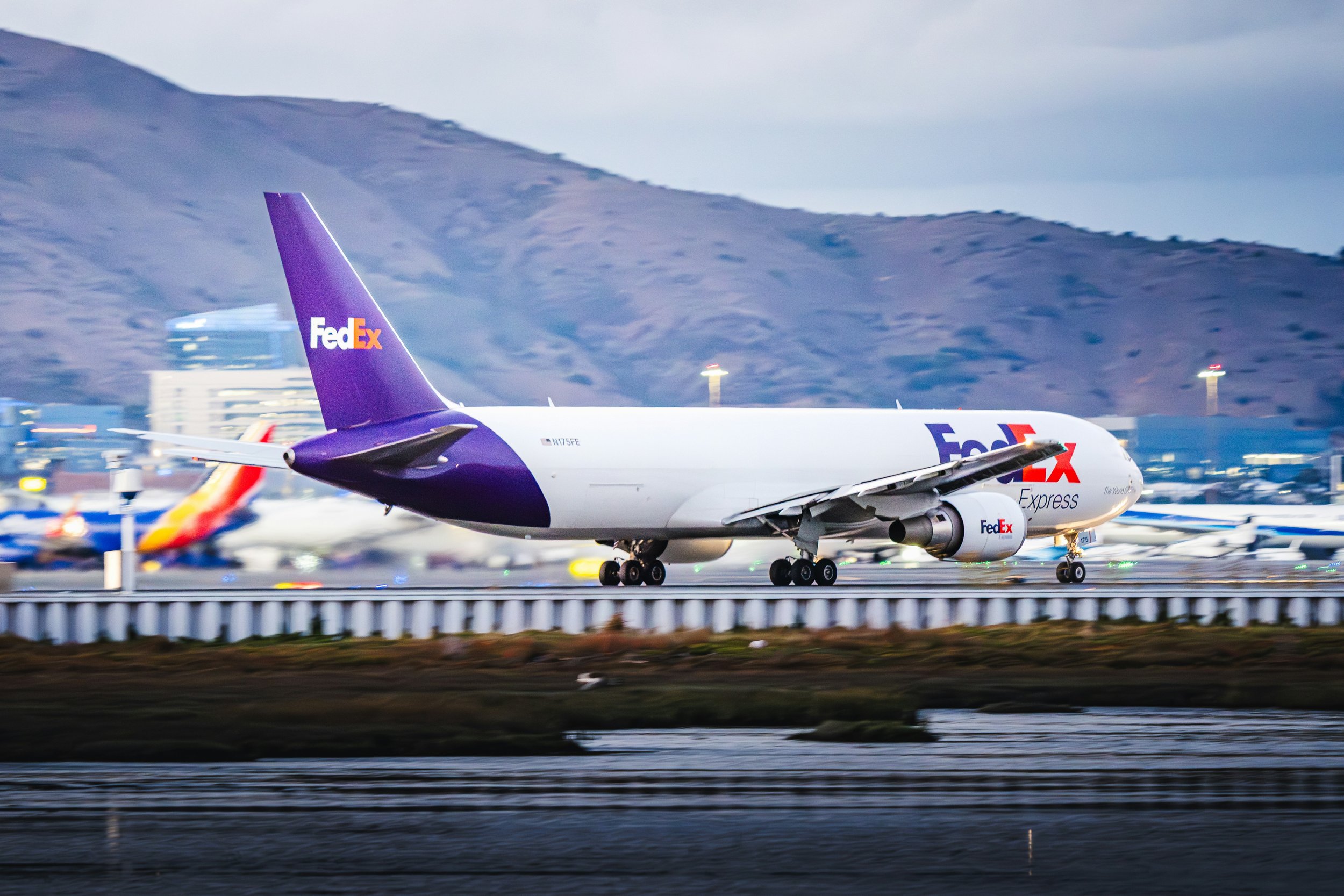 Ein FedEx-Express-Flugzeug rollt auf einer Startbahn, im Hintergrund Berge und Gebäude, während die Sonne untergeht oder aufgeht.