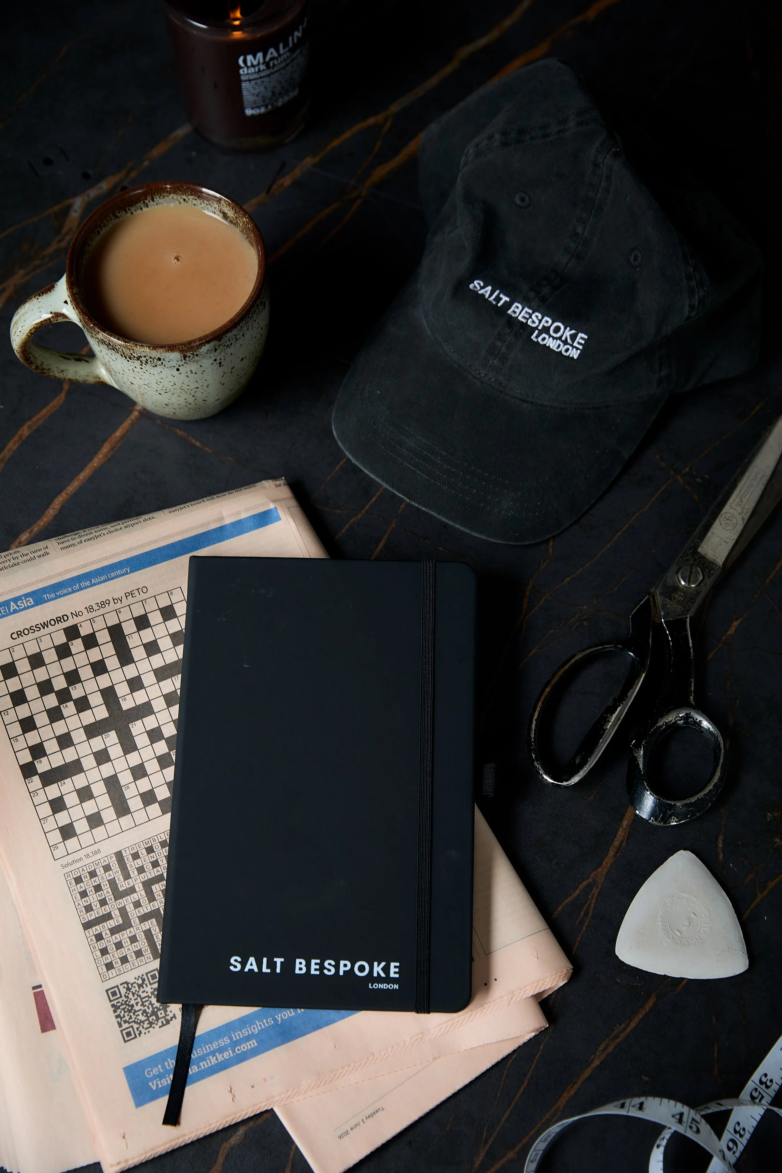 A tabletop with a cup of coffee, a black salt bespoke London notebook, a crossword puzzle, a newspaper, a pair of scissors, a tape measure, and a cream-colored object.