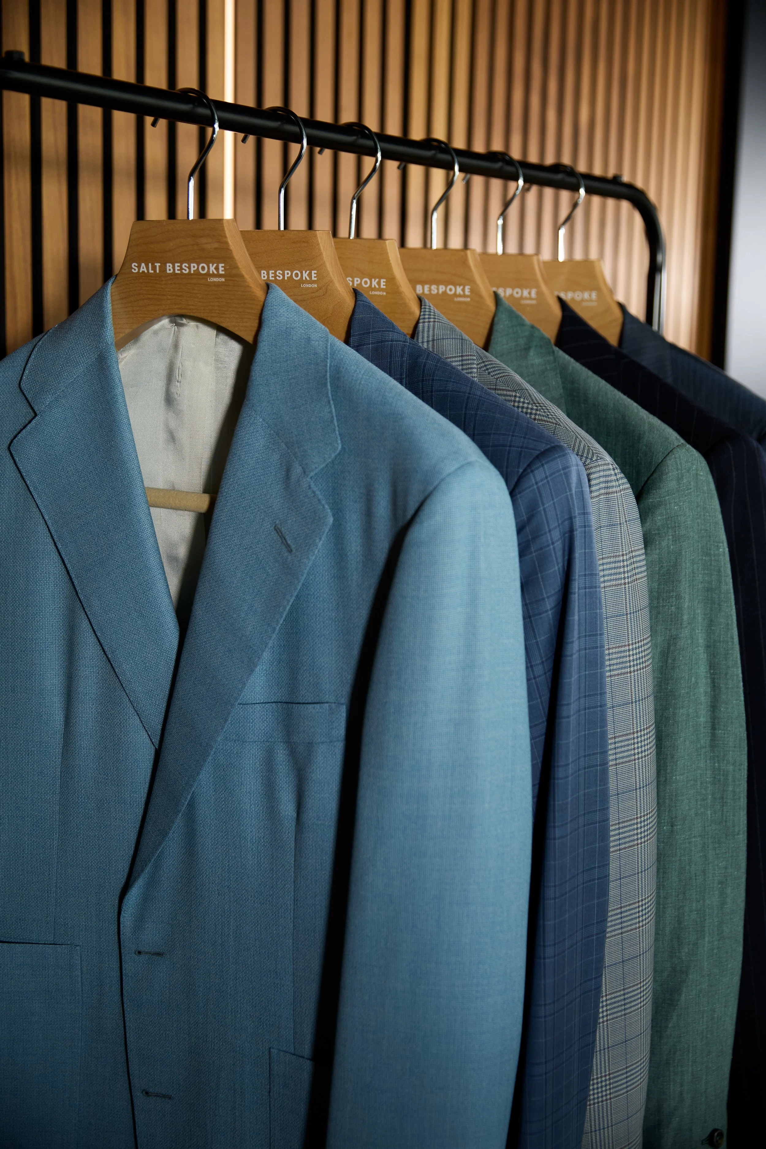 A collection of  blue, grey and green suit jackets in luxury branded fabrics hanging on wooden hangers