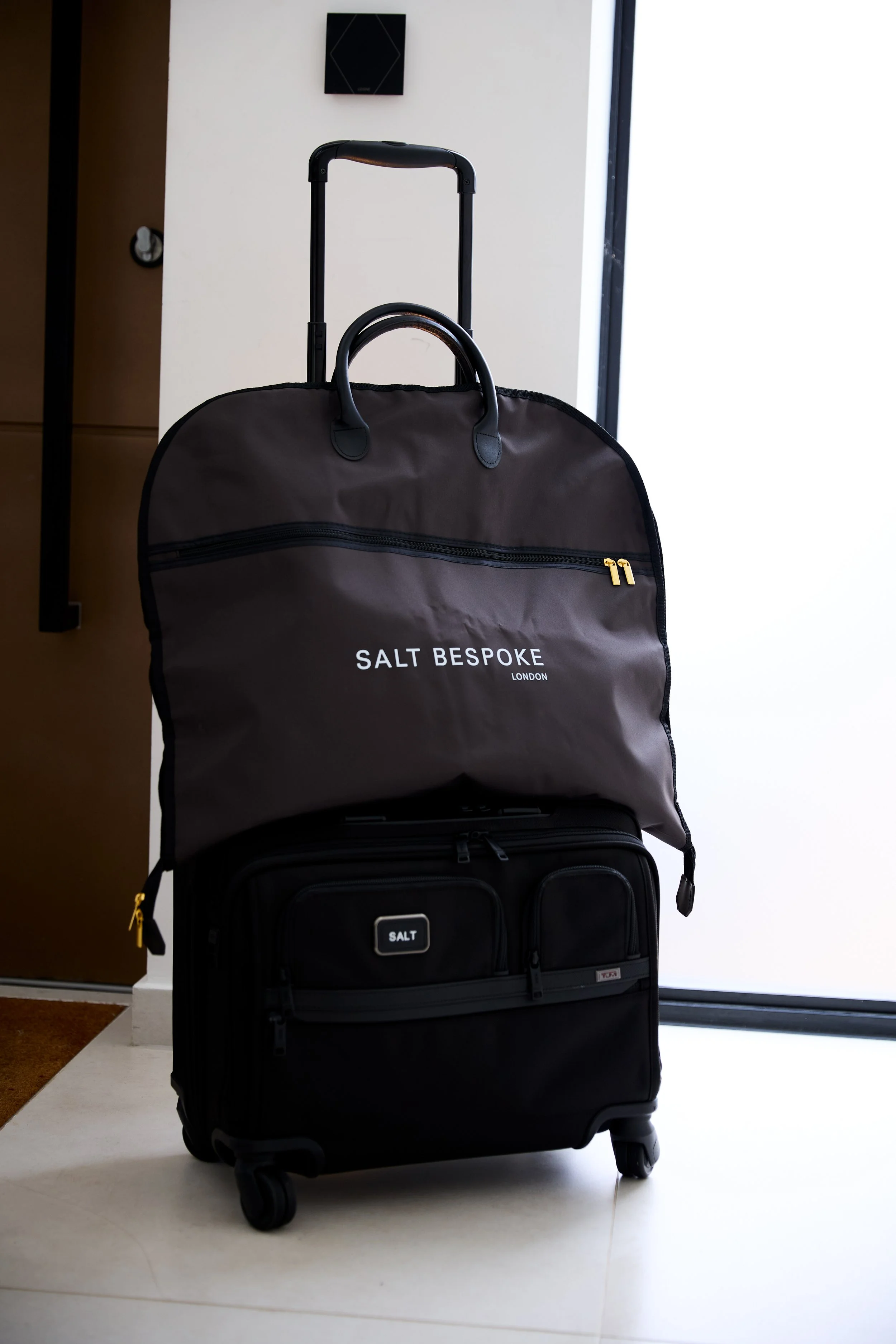 A suitcase with a suit bag on top, one black and one gray, placed near a large window