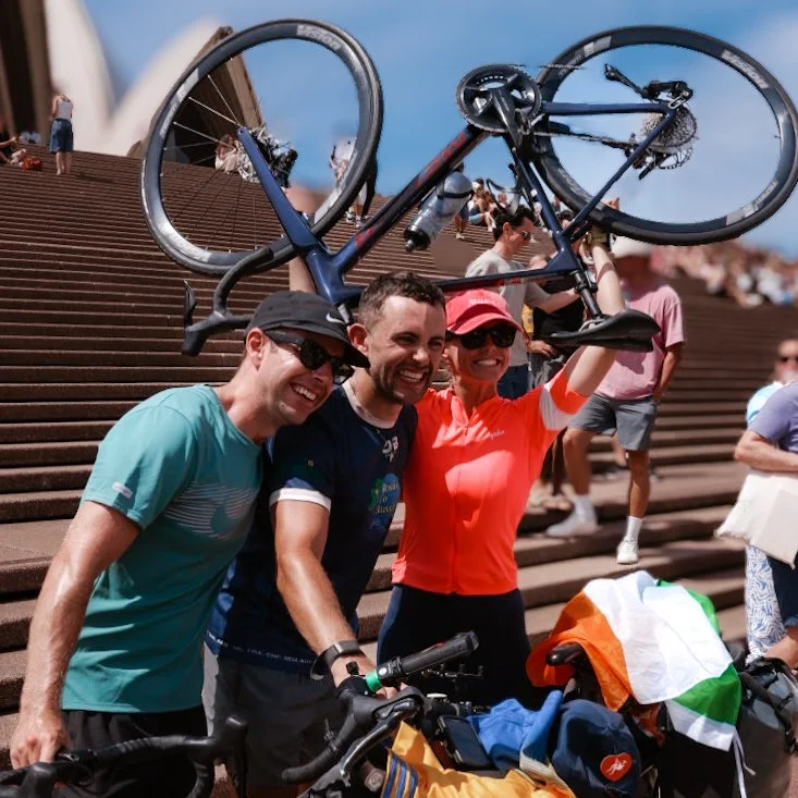 Three people smiling and posing with bikes on stairs at a scenic outdoor location, with one holding a bicycle above their head.