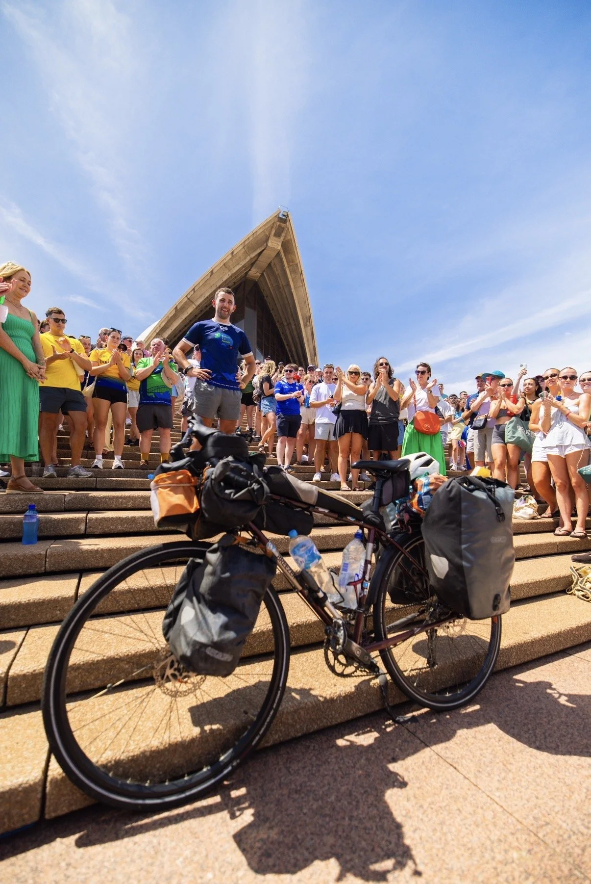 A large group of people gathered on the steps of the Sydney Opera House, with a bicycle in the foreground equipped with bags and water bottles. The crowd appears to be watching or participating in an event.