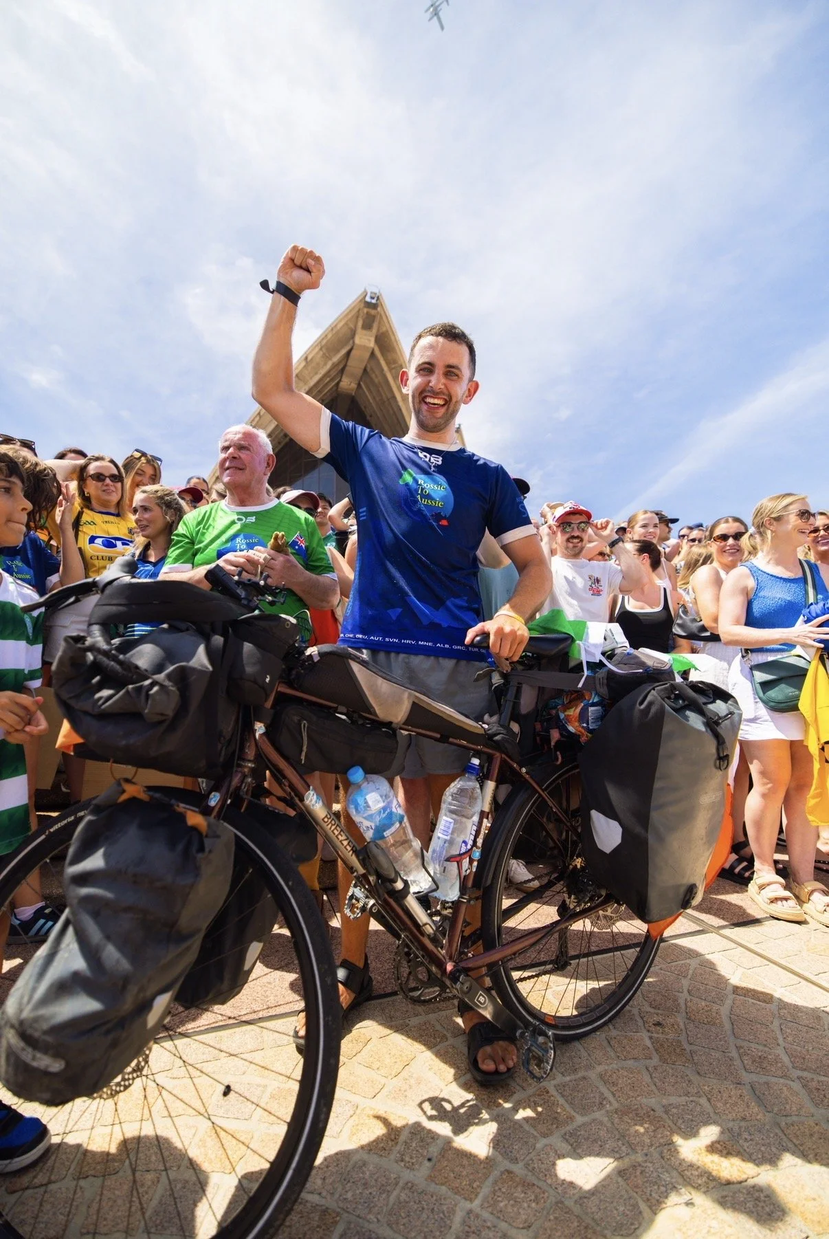 A smiling man with a bicycle, surrounded by a crowd of people, at an outdoor event with a building and plane flying overhead in the sky.