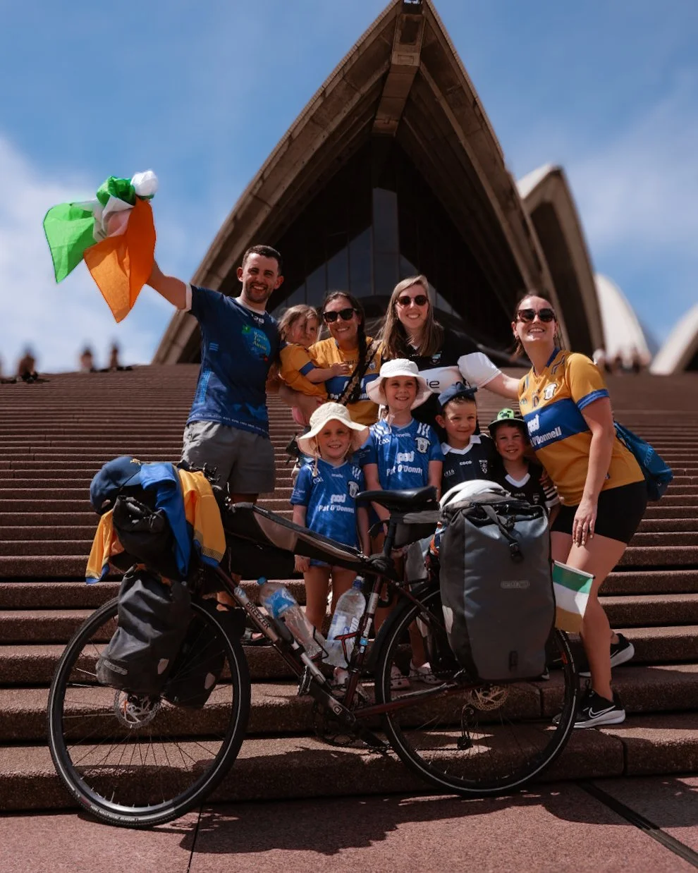 Group of people, including children and adults, posing on the steps of the Sydney Opera House, with one person holding an Irish flag and a bicycle in front.