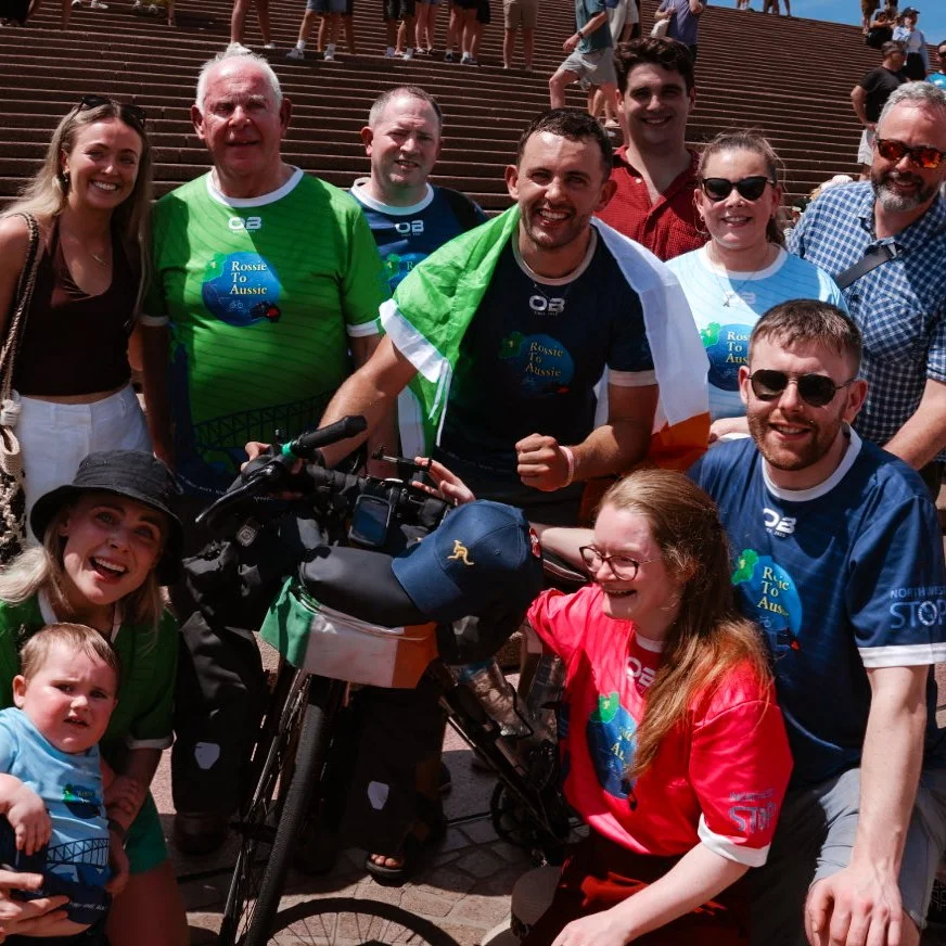 Group of people at a charity event for Rossie To Aussie, gathered around a bicycle, some wearing event t-shirts, on outdoor stairs under a clear blue sky.