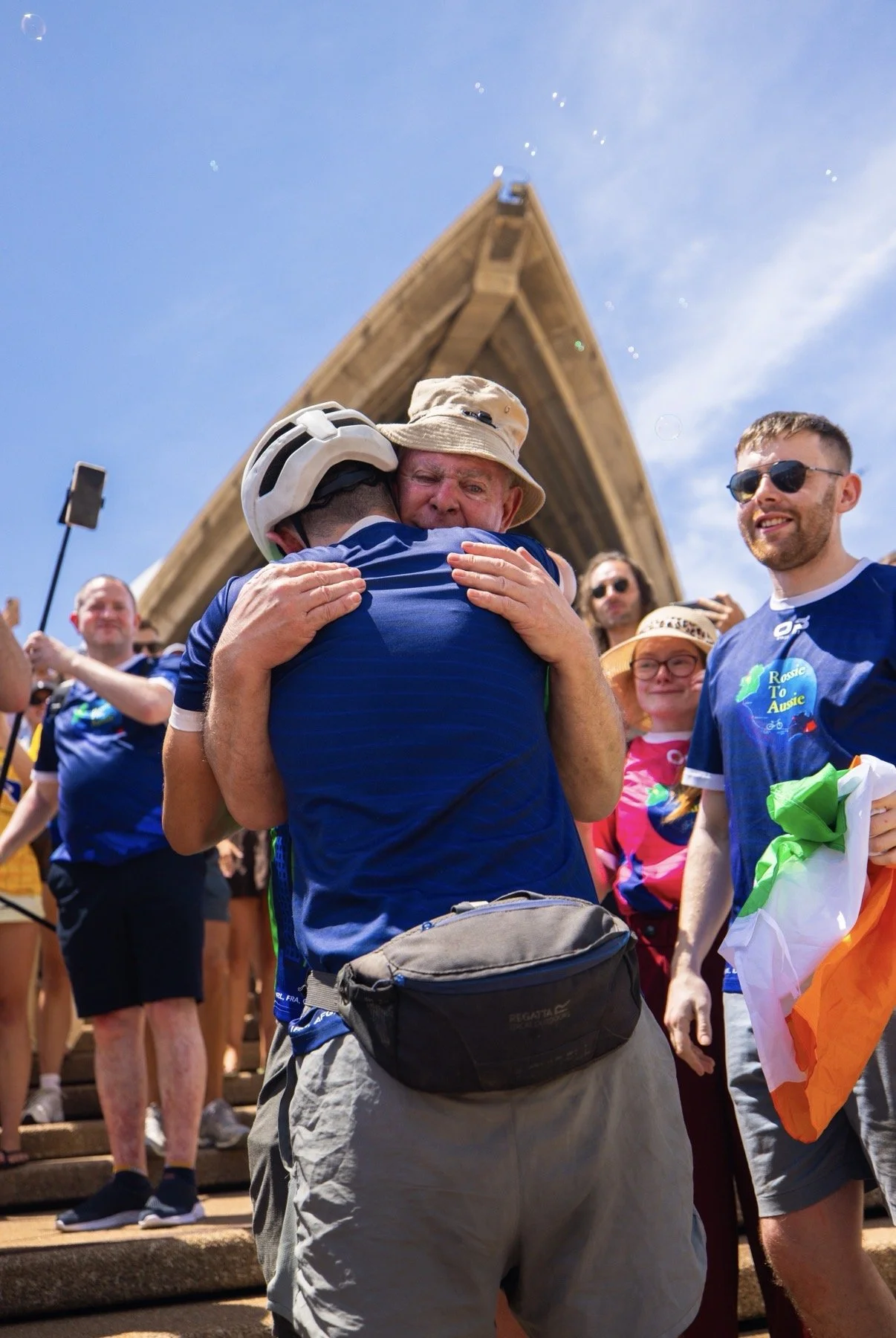 A group of people celebrating outdoors on a sunny day, with some hugging, wearing blue shirts, and one person holding an Australian flag. The background features a wooden structure and a clear blue sky.