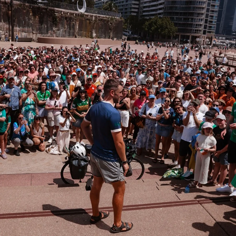 A man standing in front of a large crowd gathered outdoors in an urban area, with buildings and trees in the background.
