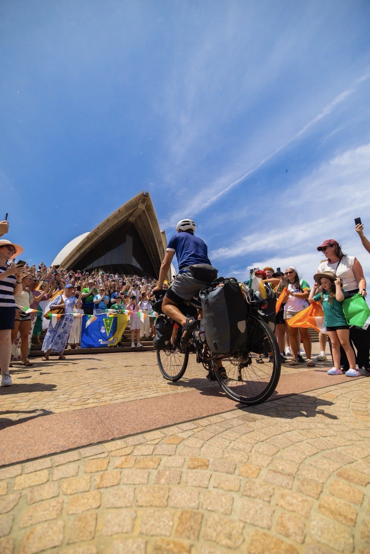A cyclist in a blue shirt and helmet riding past a crowd gathered outside the Sydney Opera House under a blue sky with faint clouds.