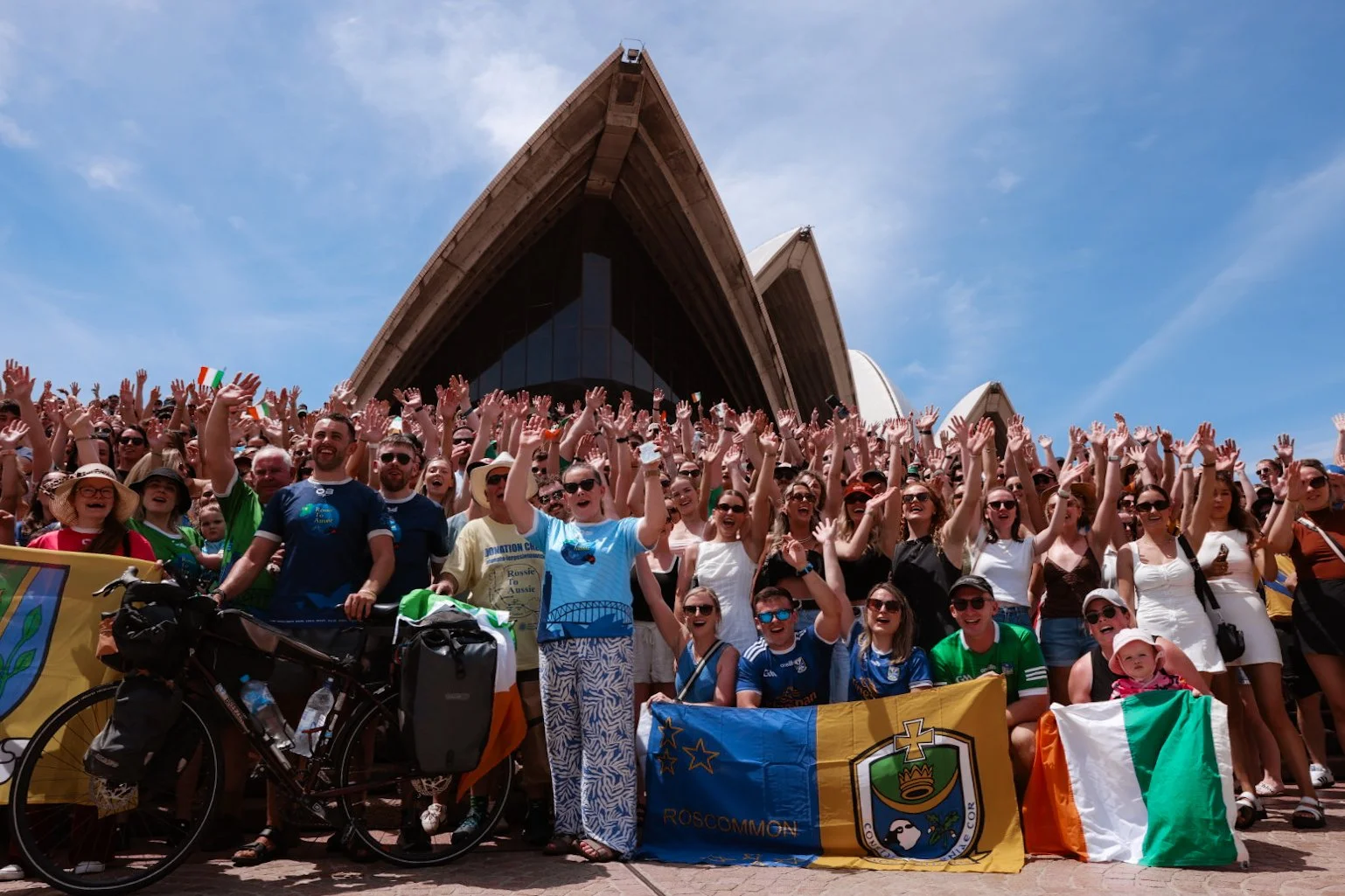 A large crowd of people gathered in front of the Sydney Opera House, celebrating and waving their hands in the air on a sunny day.