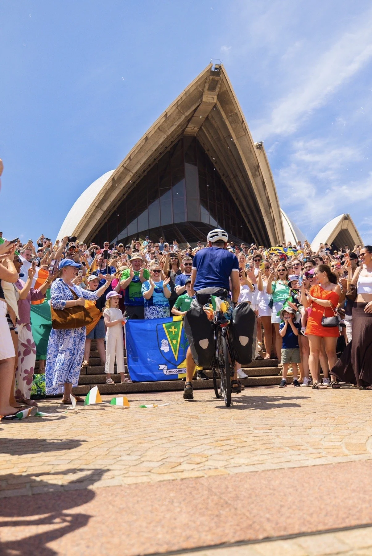 Large crowd of people gathered on the steps of the Sydney Opera House, with a cyclist in the foreground, holding a flag and facing the crowd during daytime with clear blue sky.