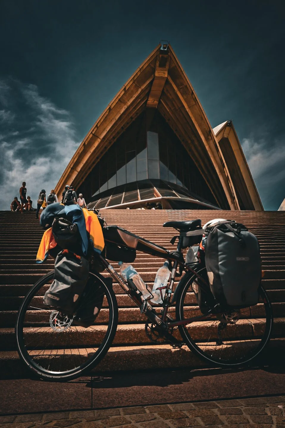A touring bicycle with gear parked on steps leading to the Sydney Opera House under a cloudy sky.
