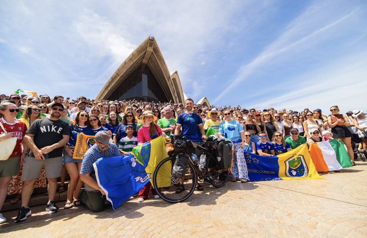 Large group of people gathered in front of the Sydney Opera House, some holding flags, with a few on bicycles, under a partly cloudy sky.