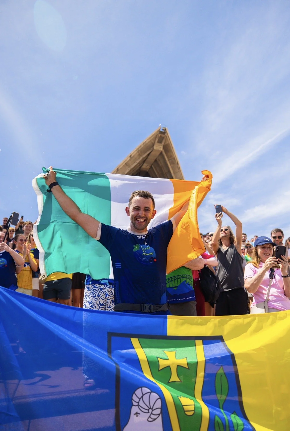 A smiling man holding an Irish flag at an outdoor event with a crowd of people around, some taking photos, and a wooden structure in the background under a clear blue sky.