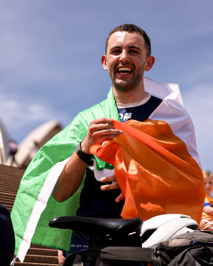 A man smiling and holding an Irish flag, with a crowd in the background, celebrating outdoors.