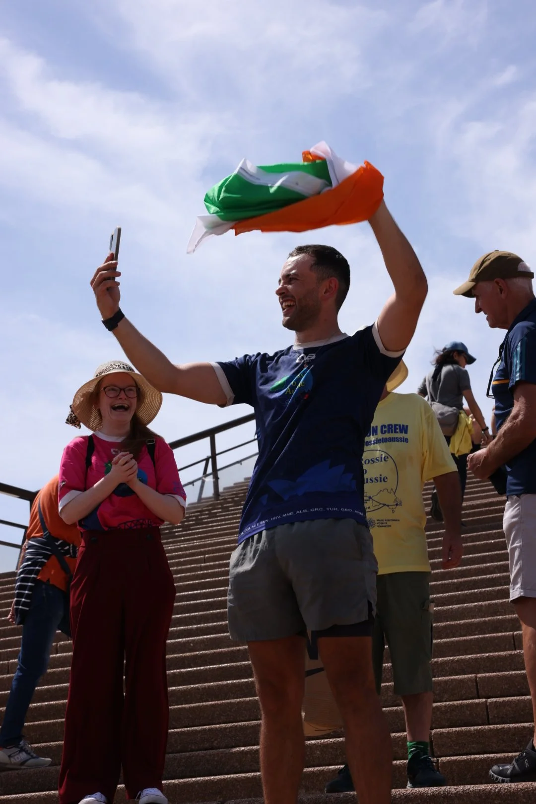 A man holding an Indian flag, smiling and taking a selfie on steps, surrounded by cheerful people, some wearing hats and casual clothing, outdoors on a sunny day.